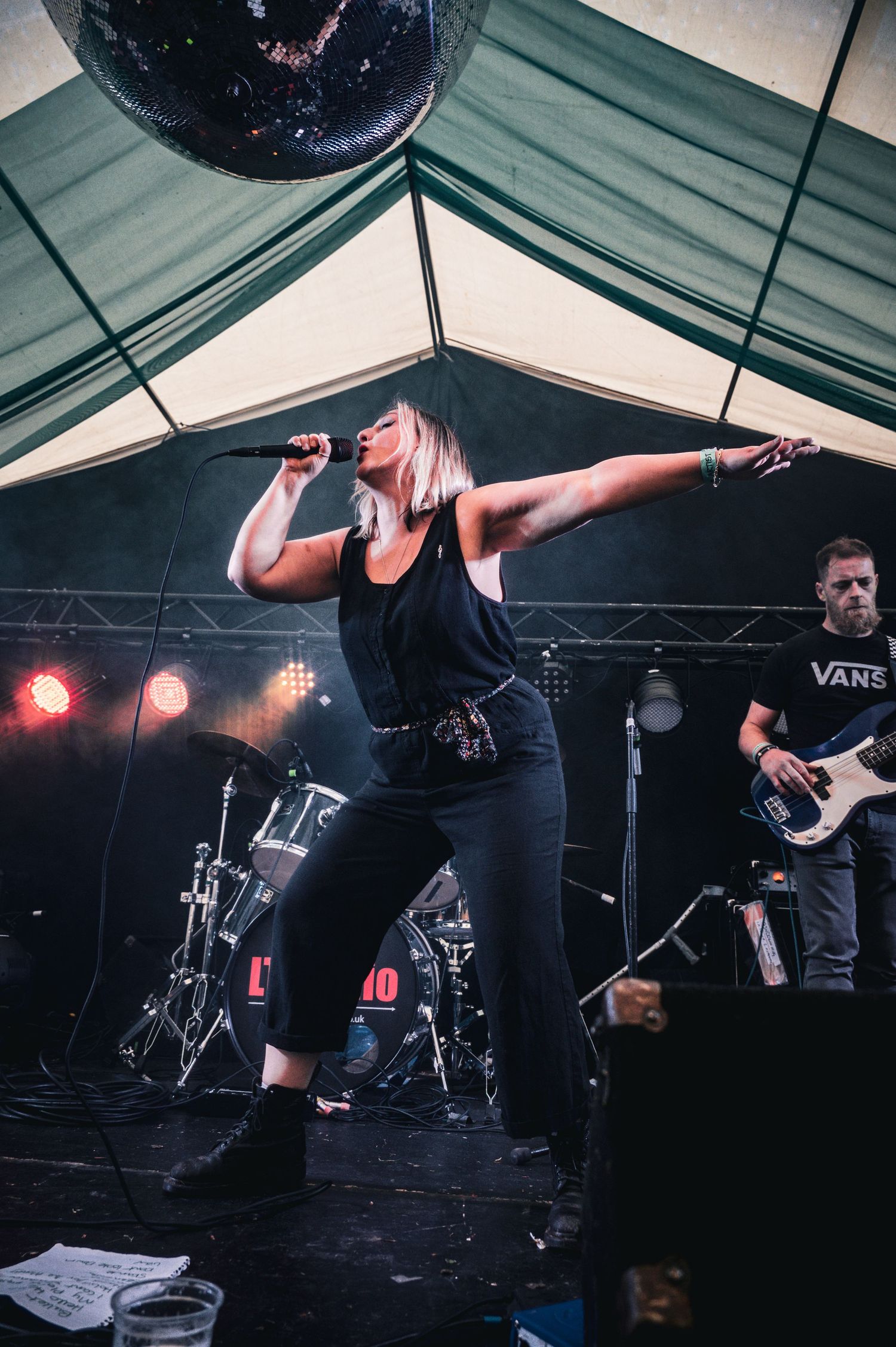 Female vocalist performing on stage with arms outstretched, guitarist in background, under tent with disco ball and stage lights.