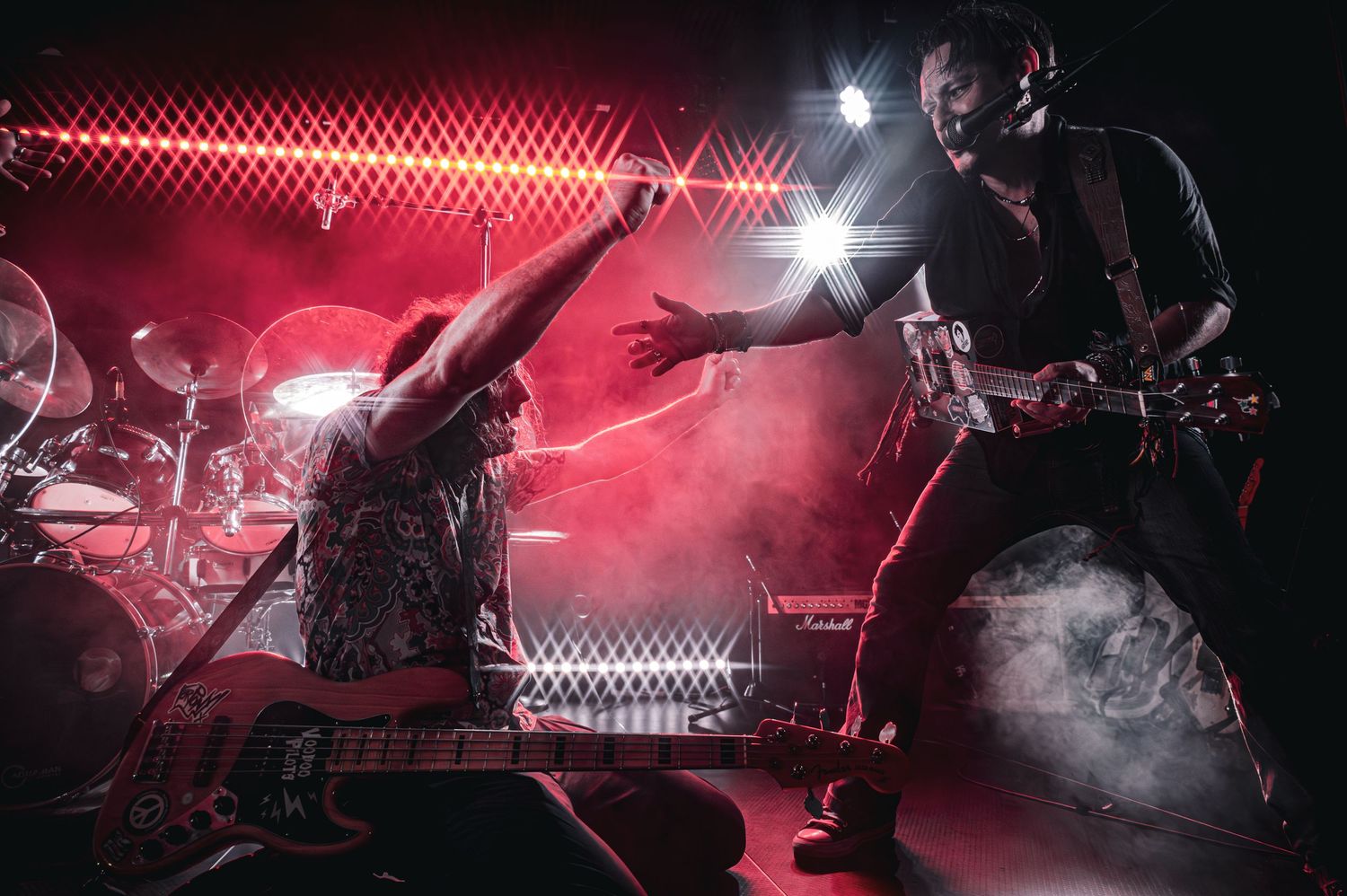 Two rock musicians perform energetically on a smoke-filled stage under dramatic red lighting at a live concert.