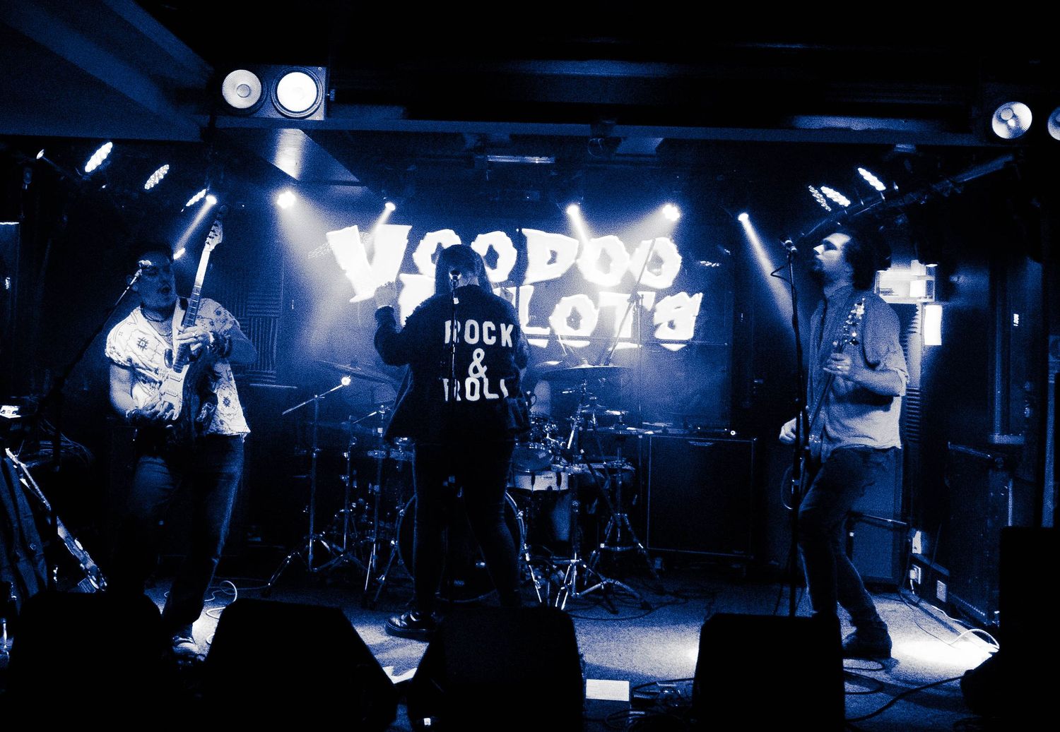 Band performing on stage with blue lighting and equipment visible in background.
