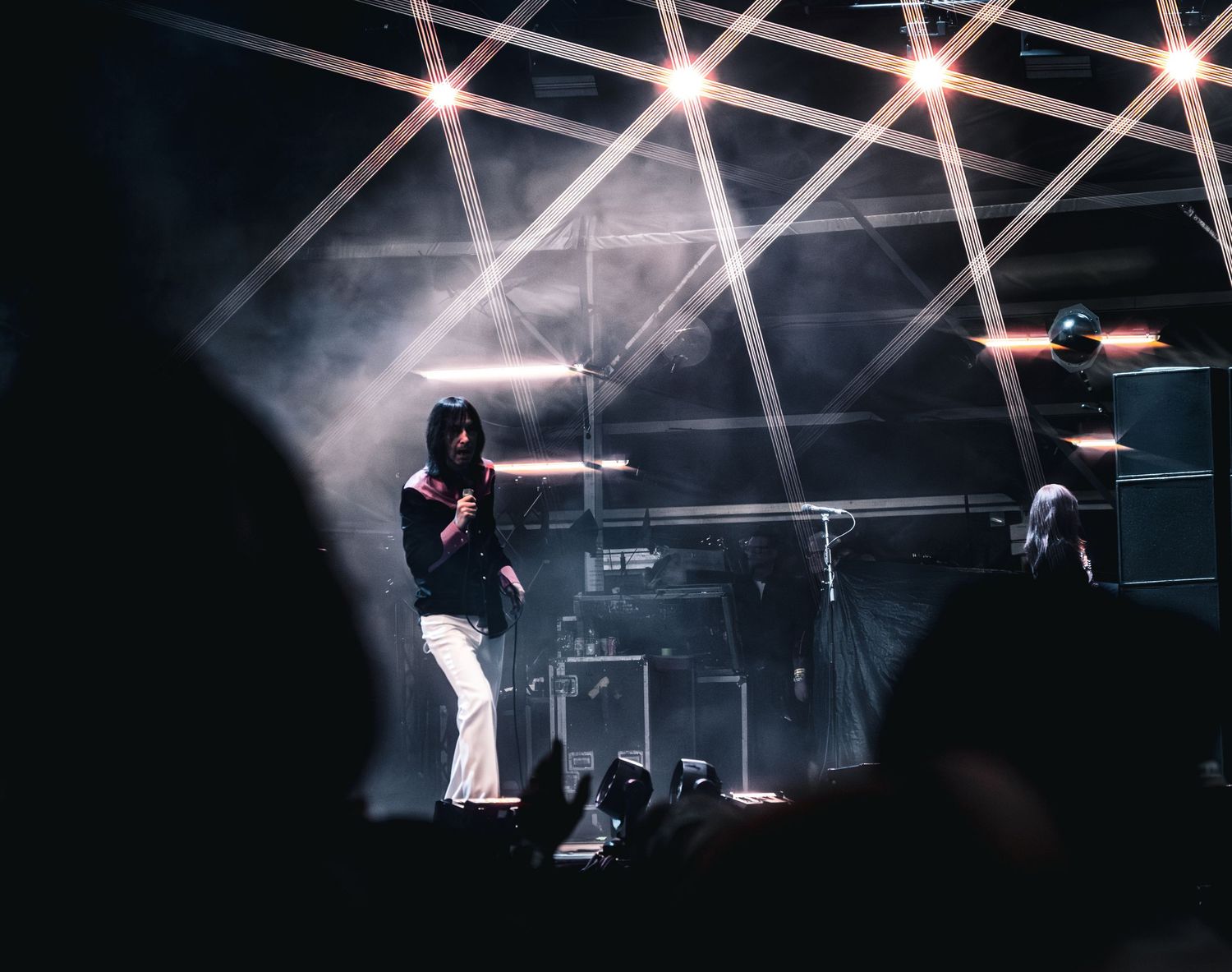 A performer sings on a dark concert stage illuminated by dramatic crossing light beams and atmospheric smoke.