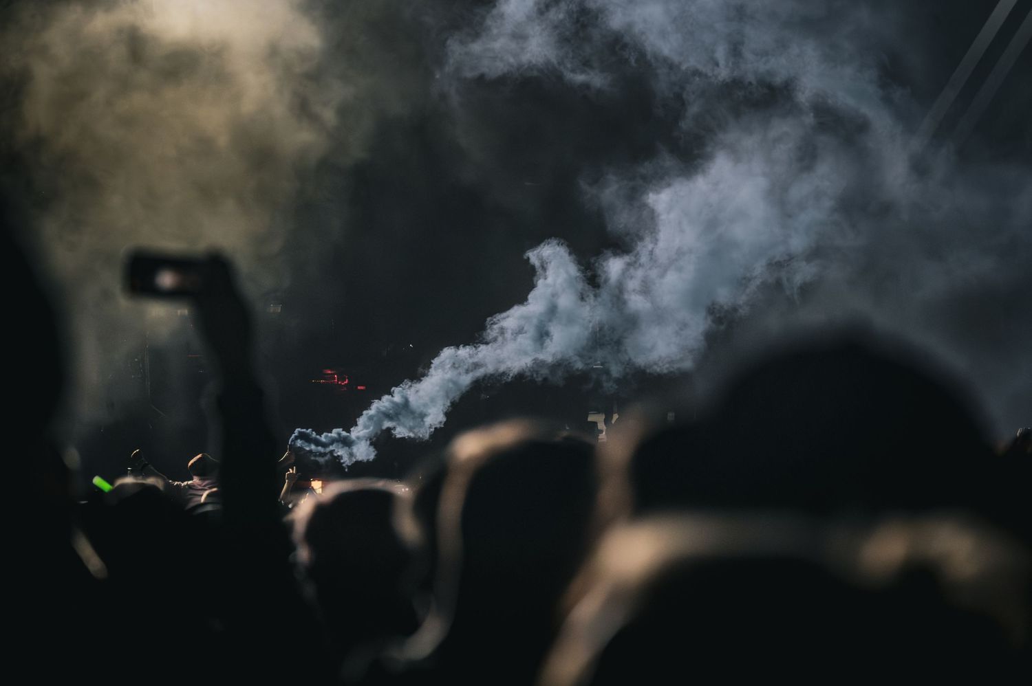 Crowd silhouettes at a dark concert venue with dramatic smoke rising through spotlights.
