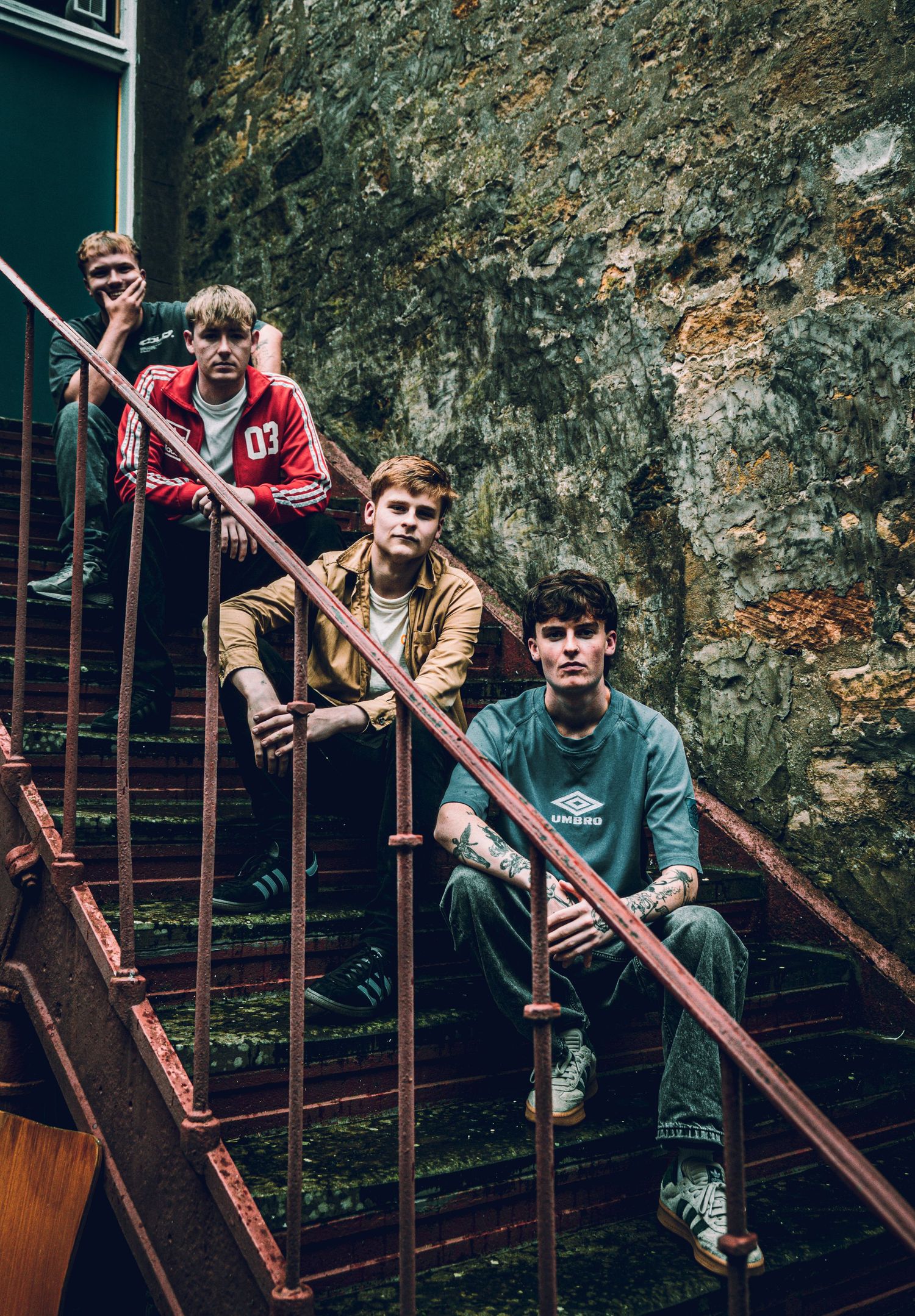 Three young men pose on rusty metal stairs against a stone wall in an urban setting.