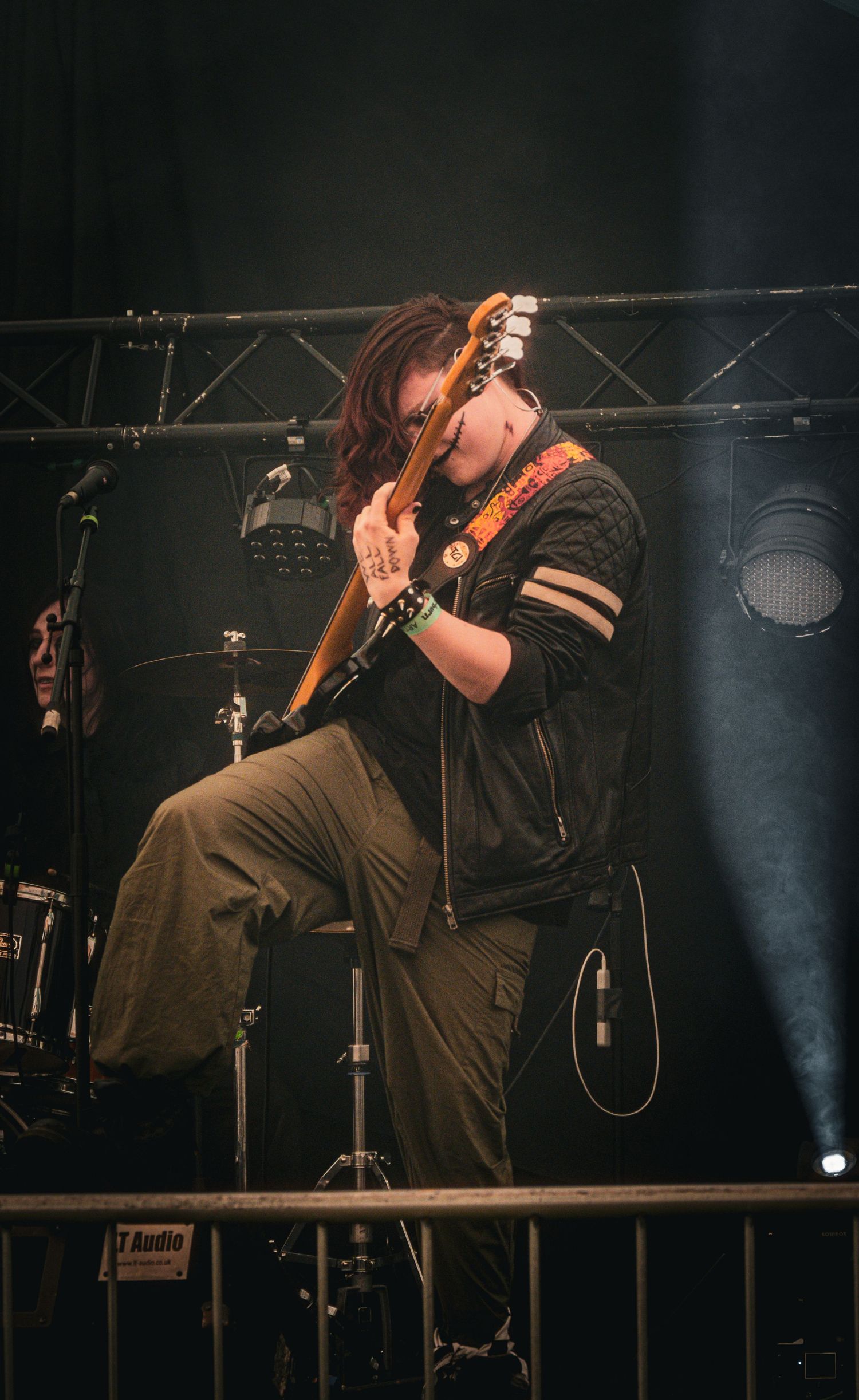 A guitarist performs on stage, passionately playing an electric guitar while seated, wearing a black jacket and olive pants.