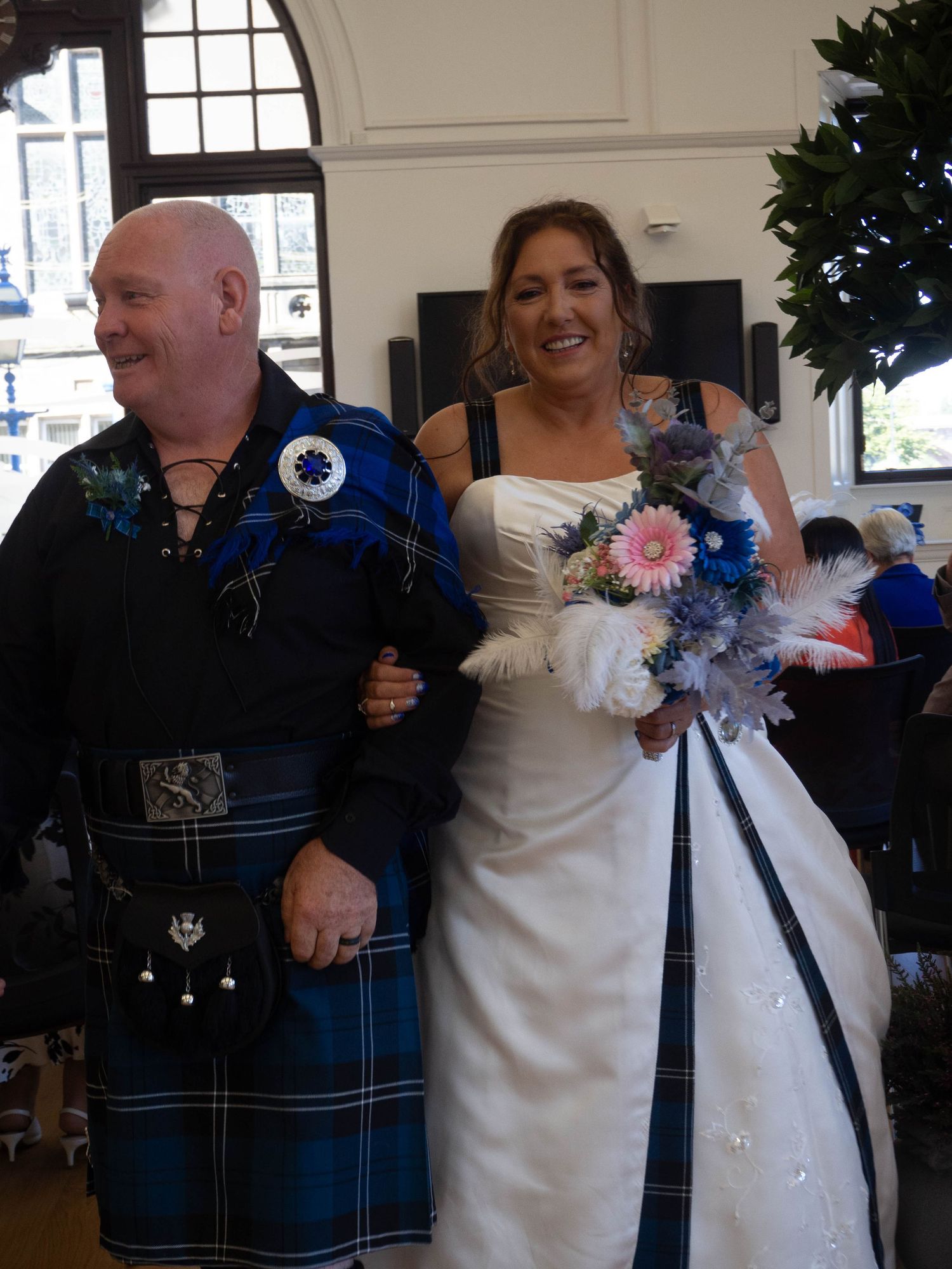 Scottish groom in blue tartan kilt walks with smiling bride in white gown holding pink and blue bouquet.
