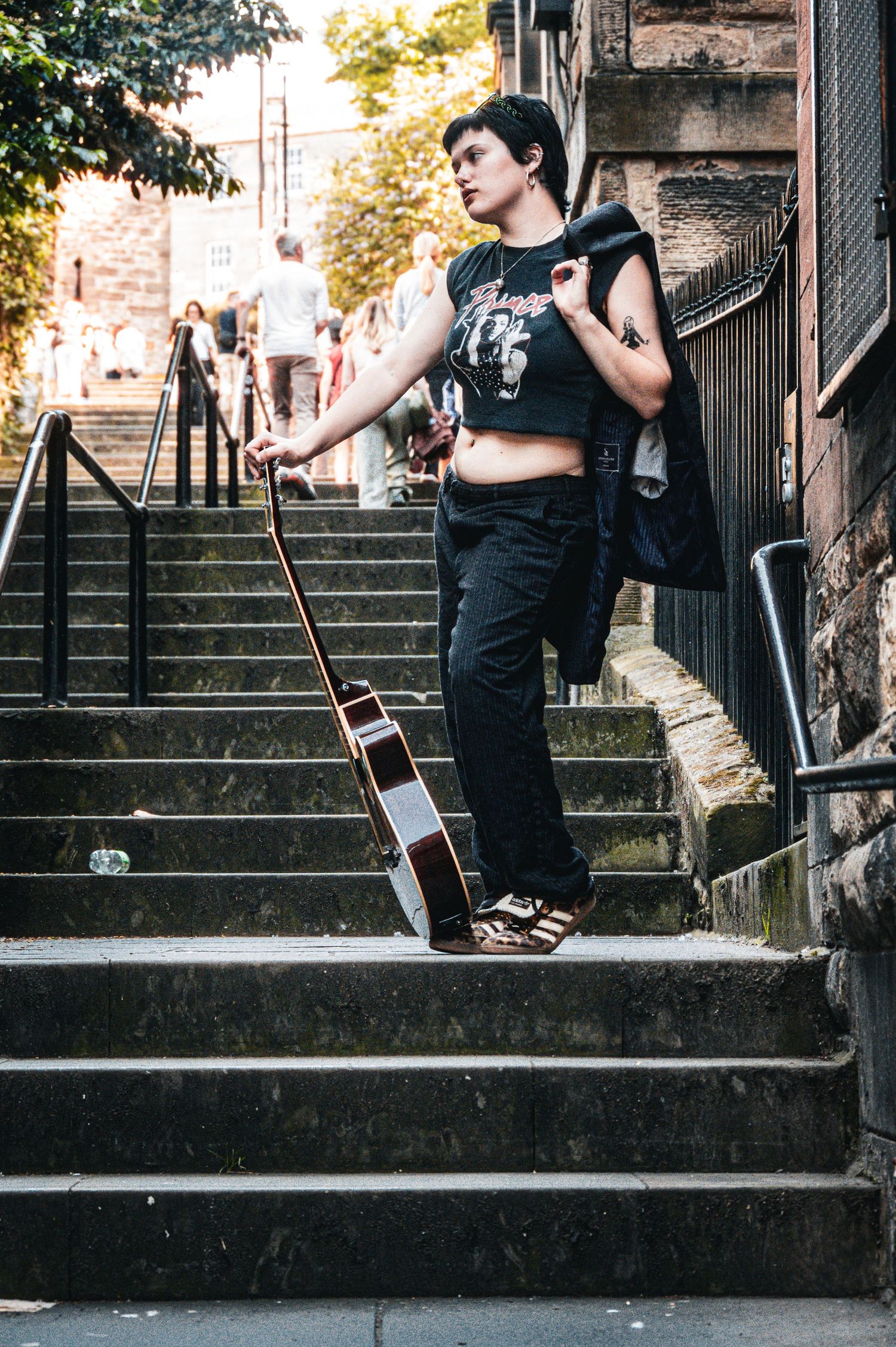 Young woman with short dark hair holding acoustic guitar on urban stone steps wearing black crop top and pants.