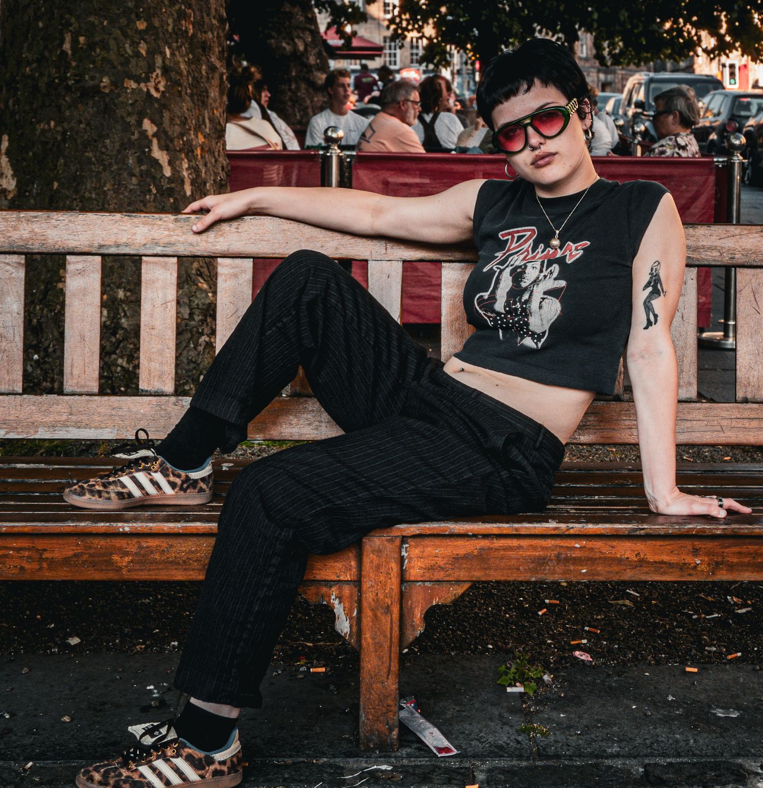 Person in black crop top and pants with red sunglasses relaxing on a wooden bench outdoors.