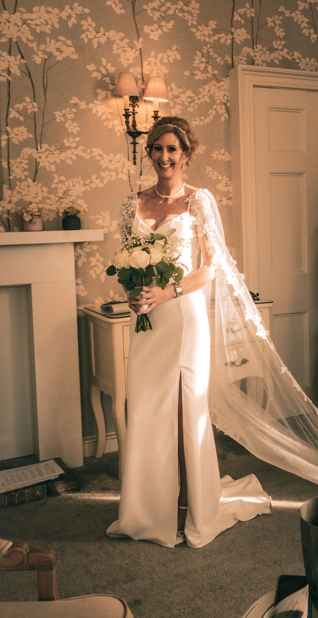 Smiling bride in a white gown with lace cape veil holding a bouquet of white roses indoors.