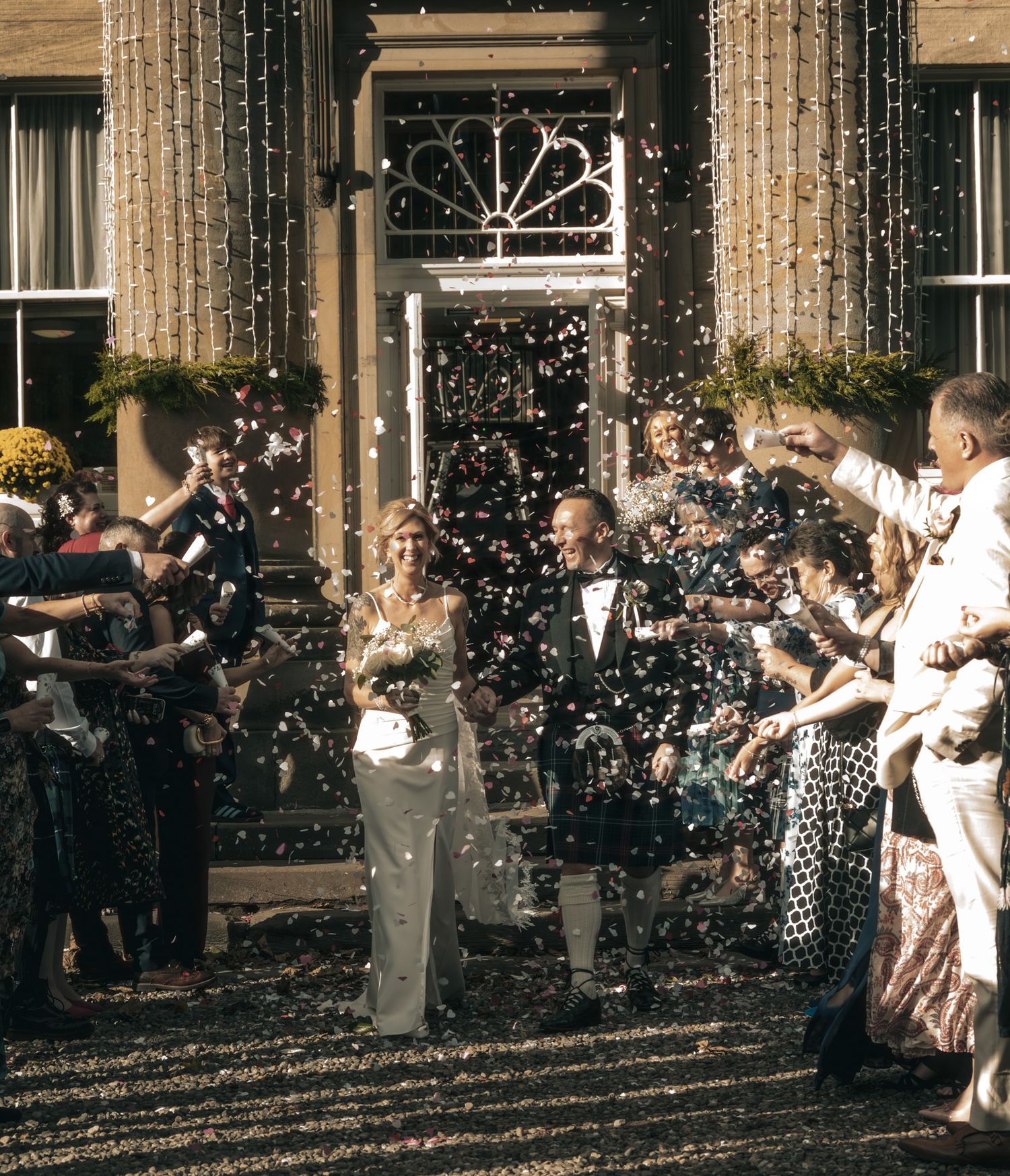 Bride and groom exit venue through confetti shower surrounded by cheering wedding guests.