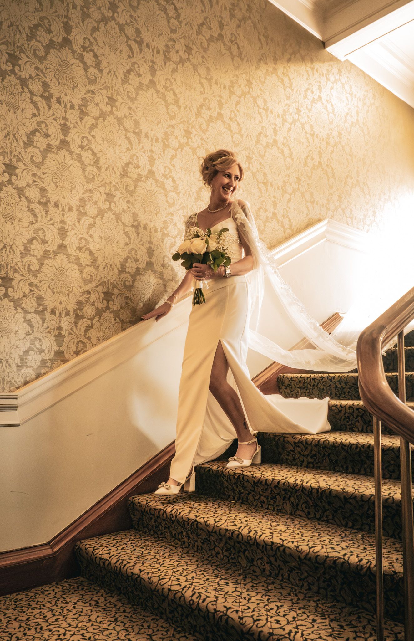 Elegant bride in a white gown holding a bouquet descends a grand staircase with ornate wallpaper at a wedding venue.