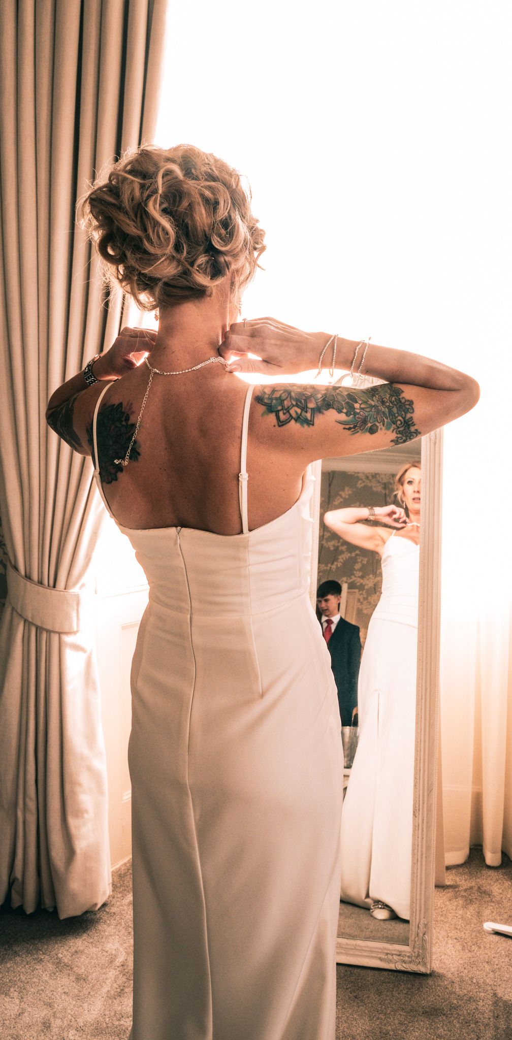 Tattooed bride in a white backless wedding dress adjusting her necklace in front of a mirror.