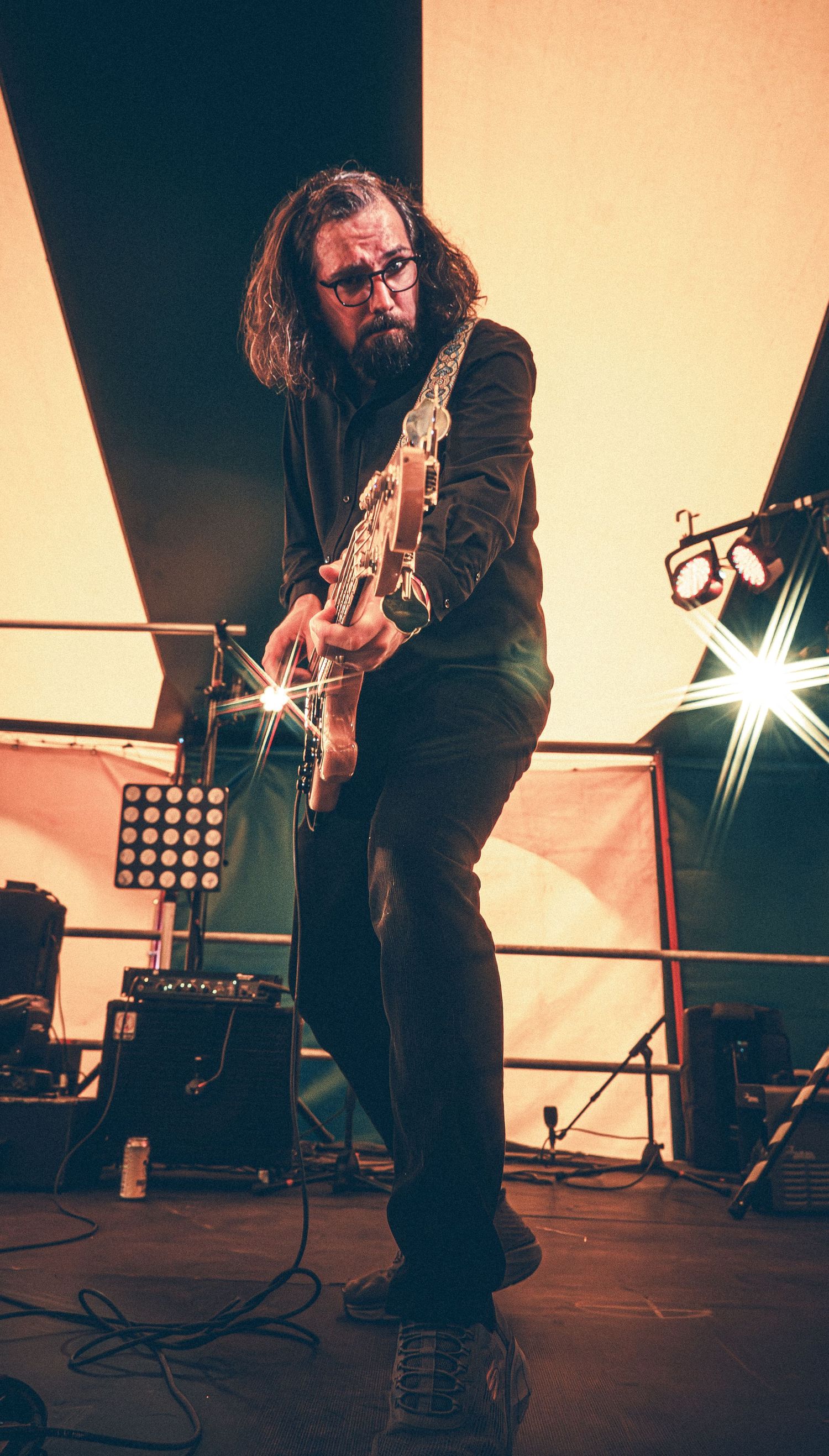 Bearded guitarist with long hair and glasses performing on stage in black outfit under bright white stage lights.