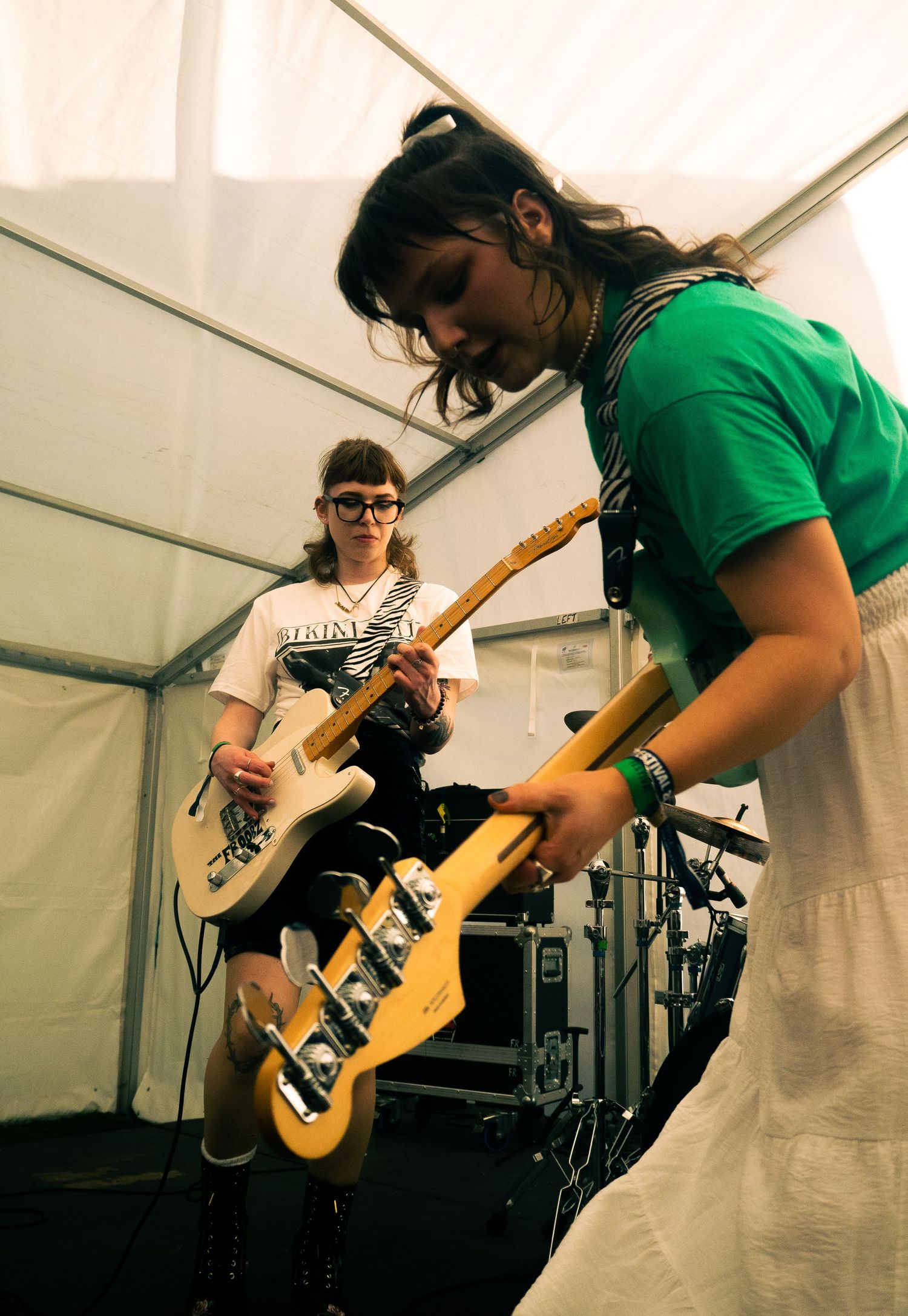 Two musicians perform on stage under a white tent, playing bass guitars with indie rock energy.