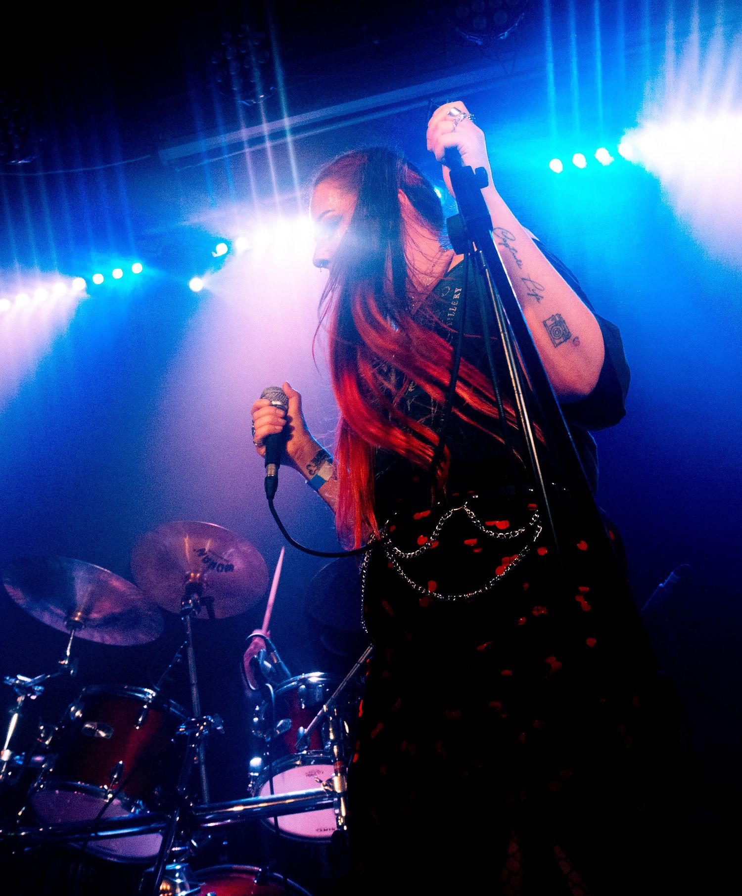 Concert performer sings on stage with dramatic blue lighting and drum kit visible in background.