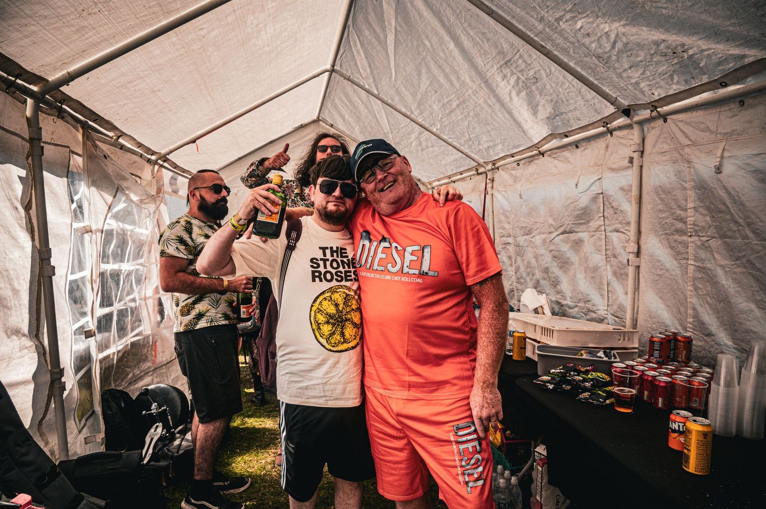 Two men pose under a tent at an outdoor festival, one wearing a The Stone Roses shirt, surrounded by colorful cans.