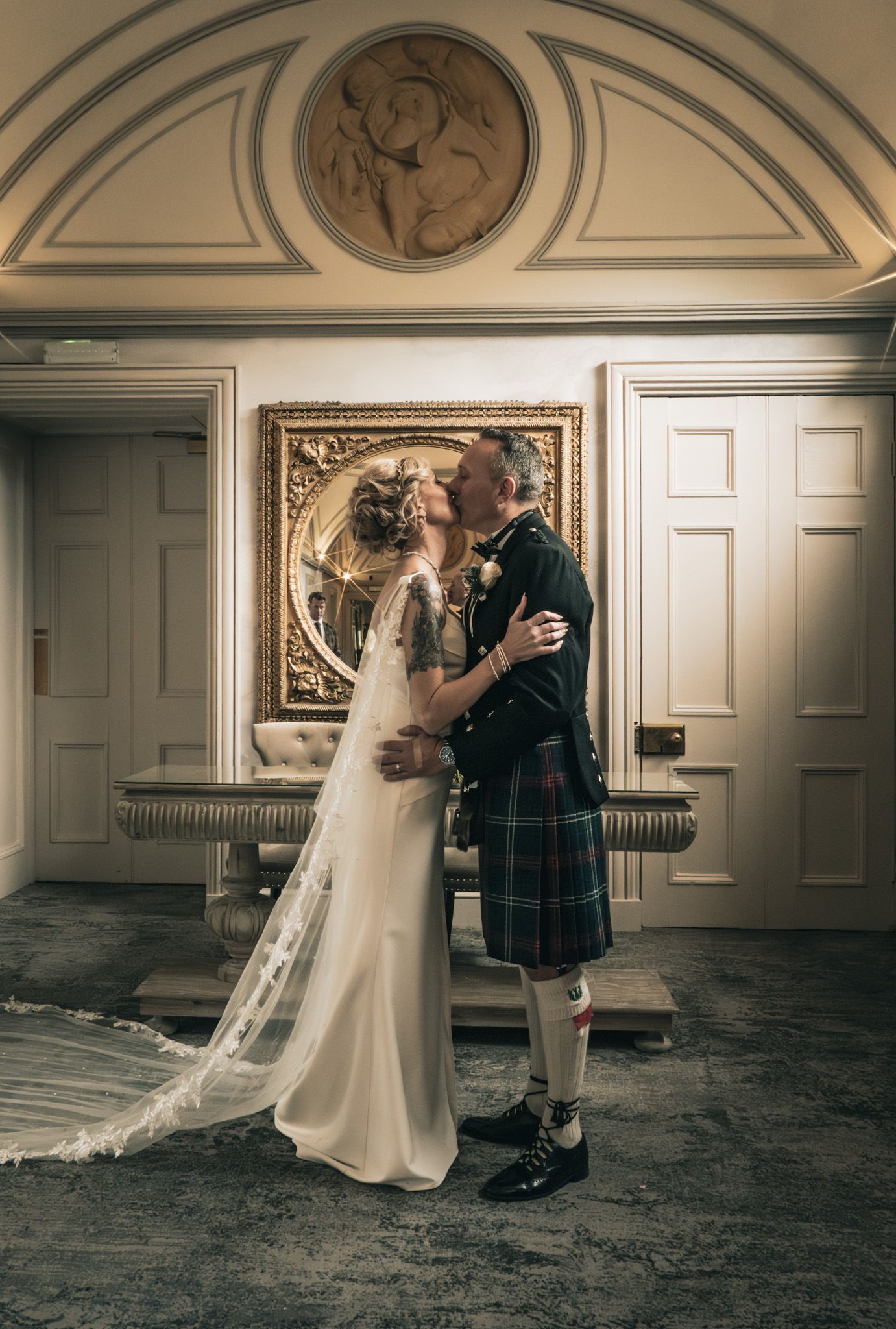 Bride and groom share a kiss indoors, groom wearing traditional Scottish tartan kilt.