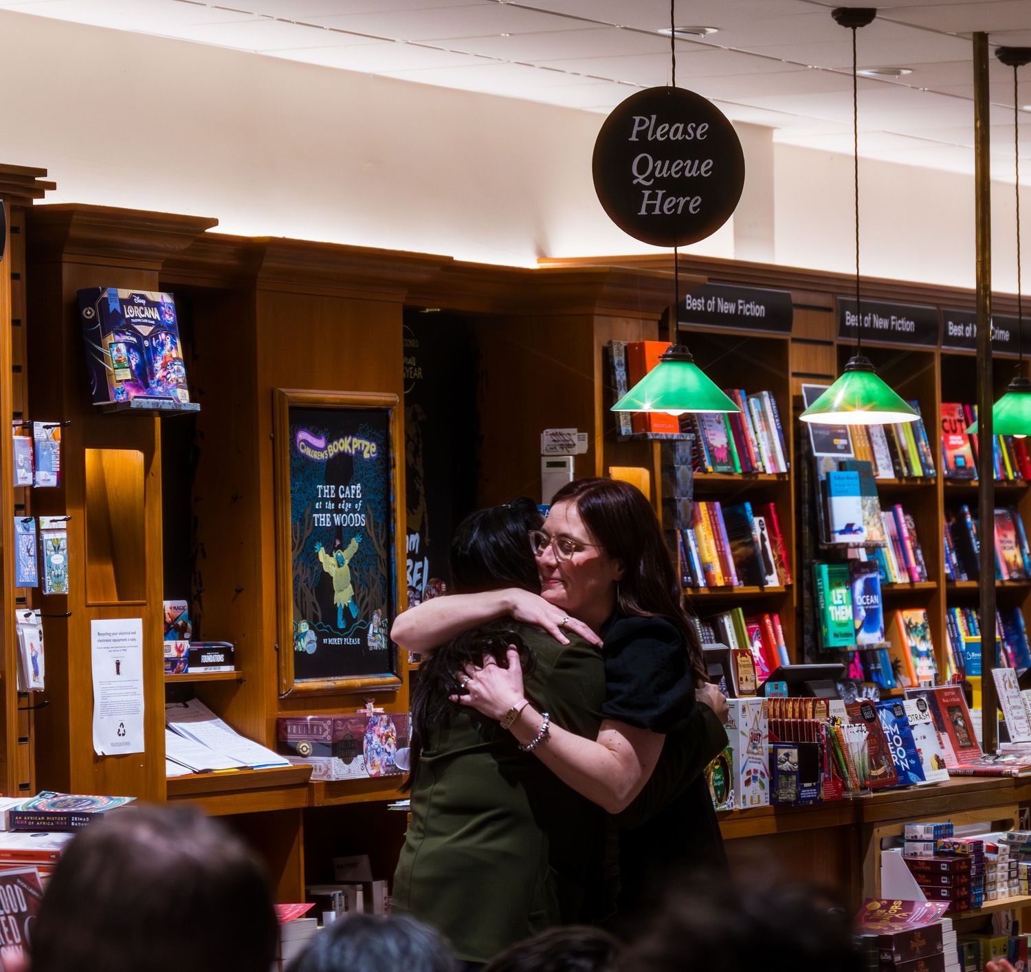 Two women sharing a warm hug in a bookstore near a Please Queue Here sign, surrounded by shelves of books.