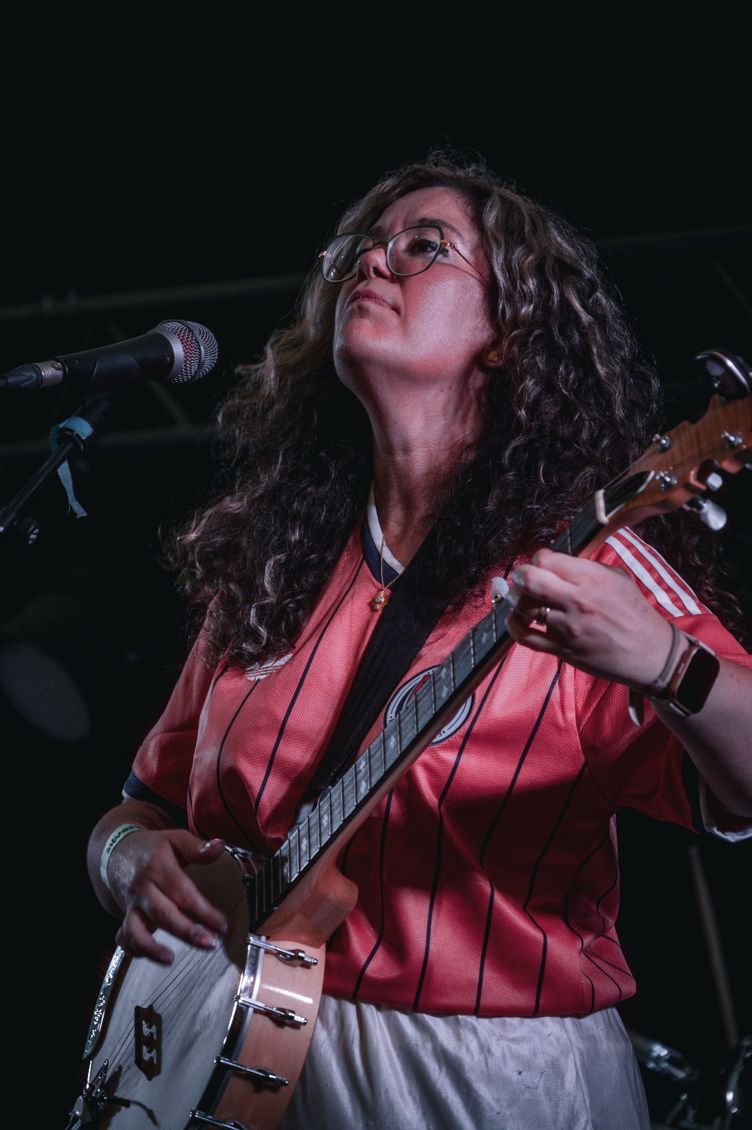 Curly-haired woman in red striped jersey playing guitar on stage near microphone under dark concert lighting.