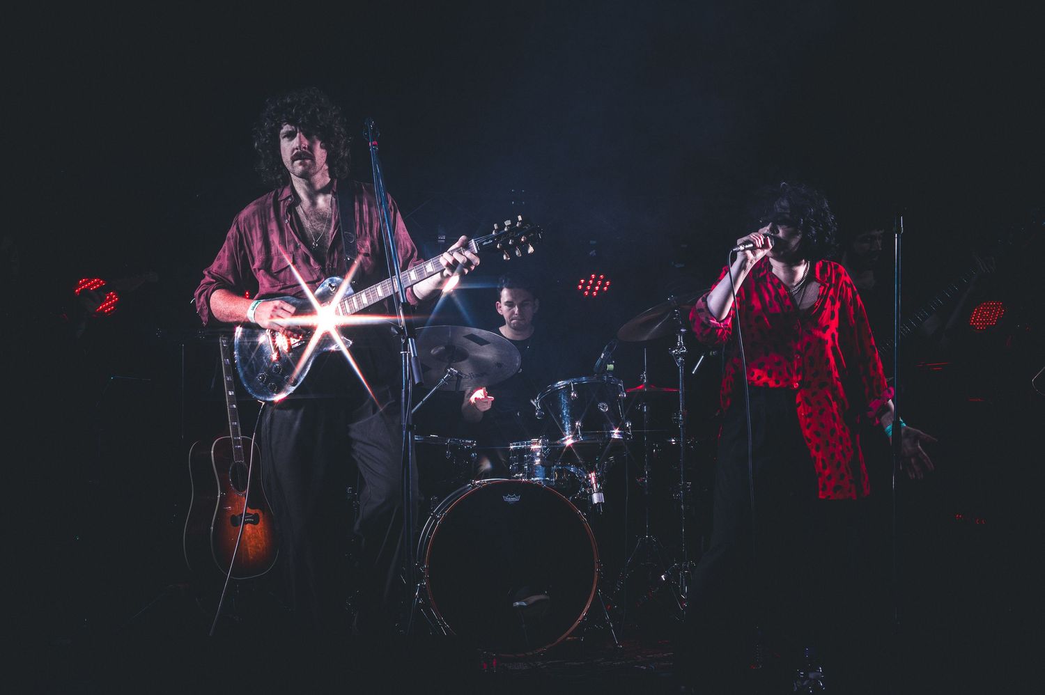 Rock band performing live on stage with guitarist, drummer, and vocalist in dramatic red lighting.