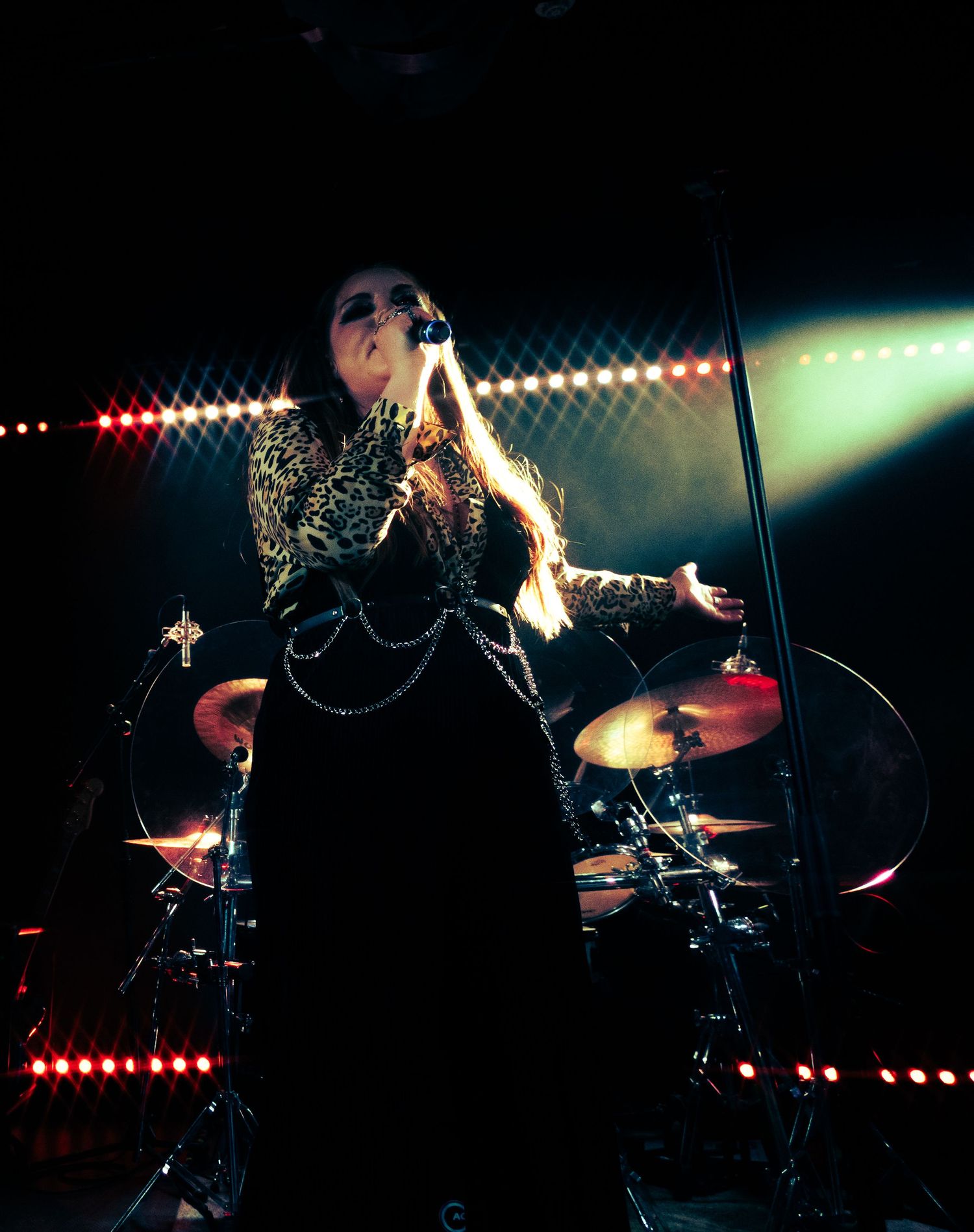 A performer in a sparkly black outfit singing on stage with dramatic lighting and a drum kit visible in the background.