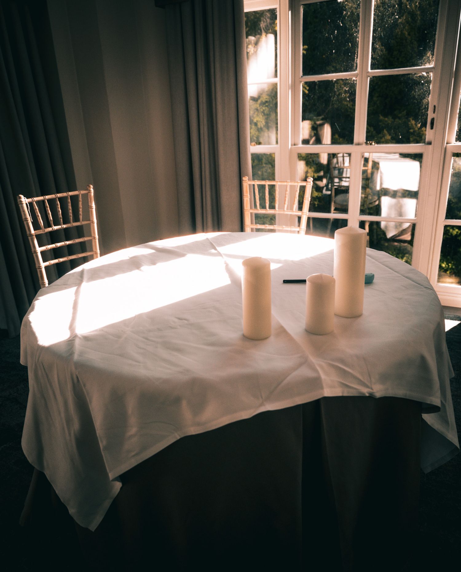 Elegant round table with white tablecloth and three pillar candles near sunlit French windows with gold Chiavari chairs.