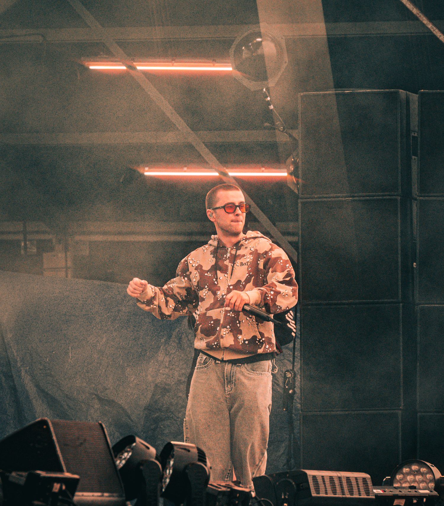 A performer in a patterned hoodie and glasses stands on a foggy stage under red neon lights with speakers behind him.