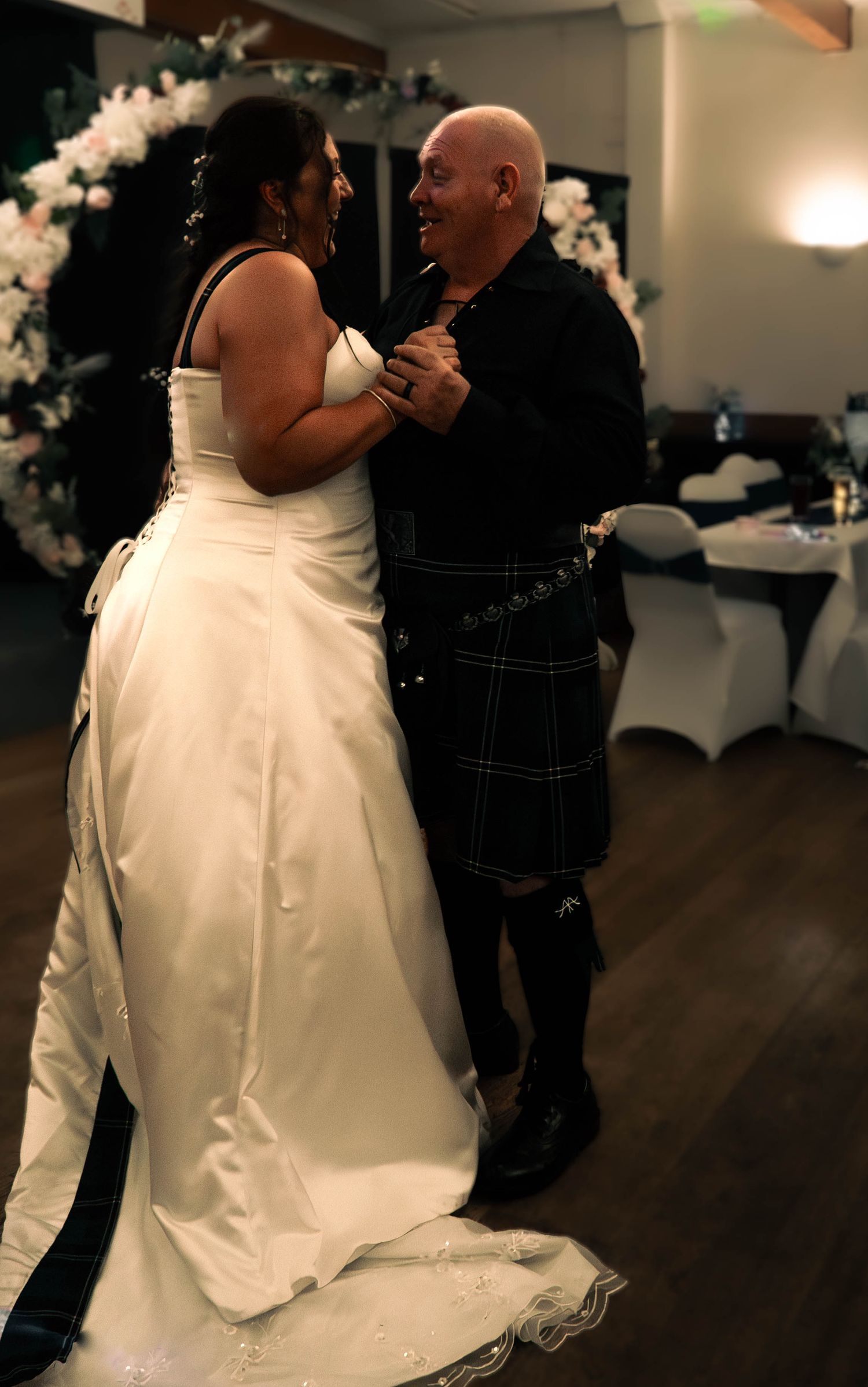 Newlywed couple shares first dance, bride in white gown and groom in dark tartan kilt, smiling joyfully.