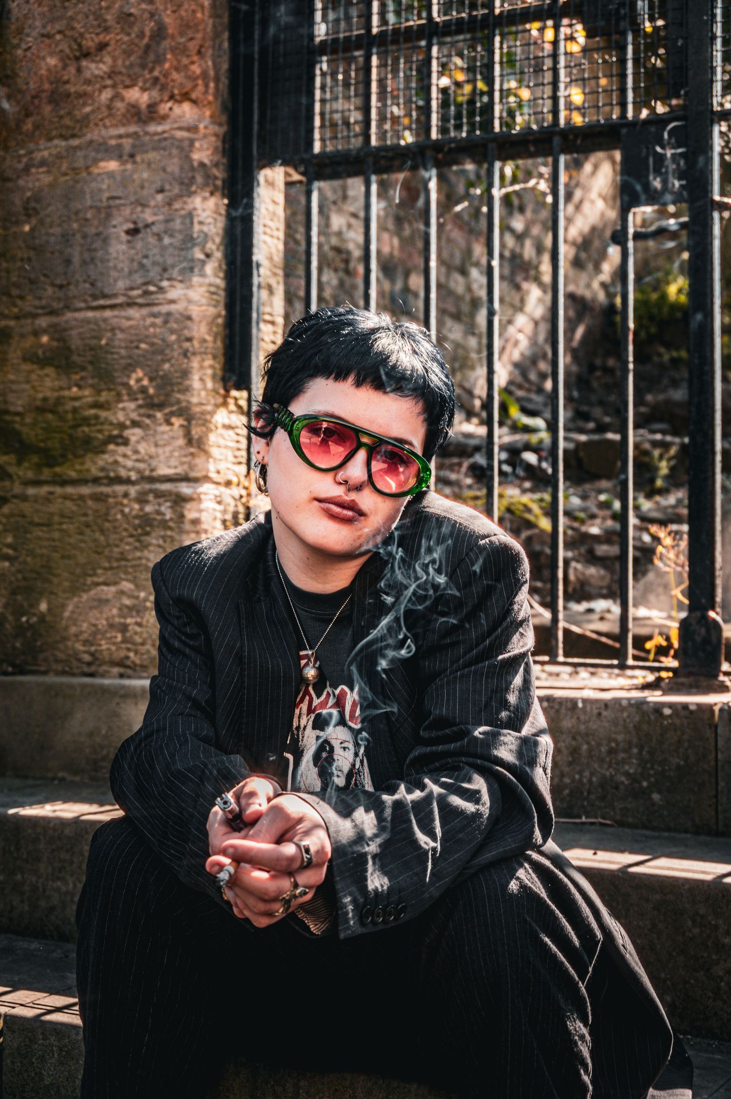 Person with short dark hair and red-green sunglasses smoking while seated on stone steps outside.