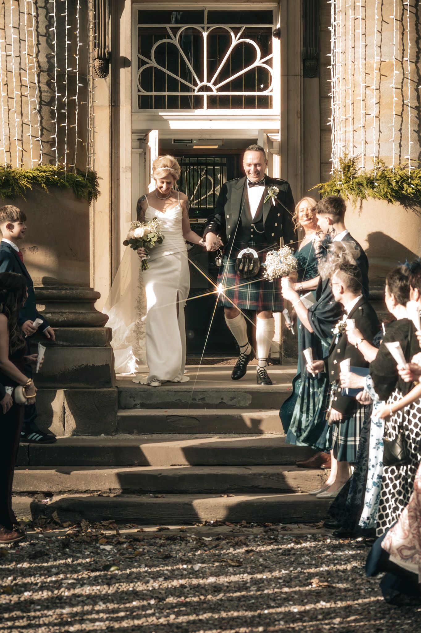 Newlyweds in white gown and Scottish kilt exit church steps as guests celebrate with confetti streamers.