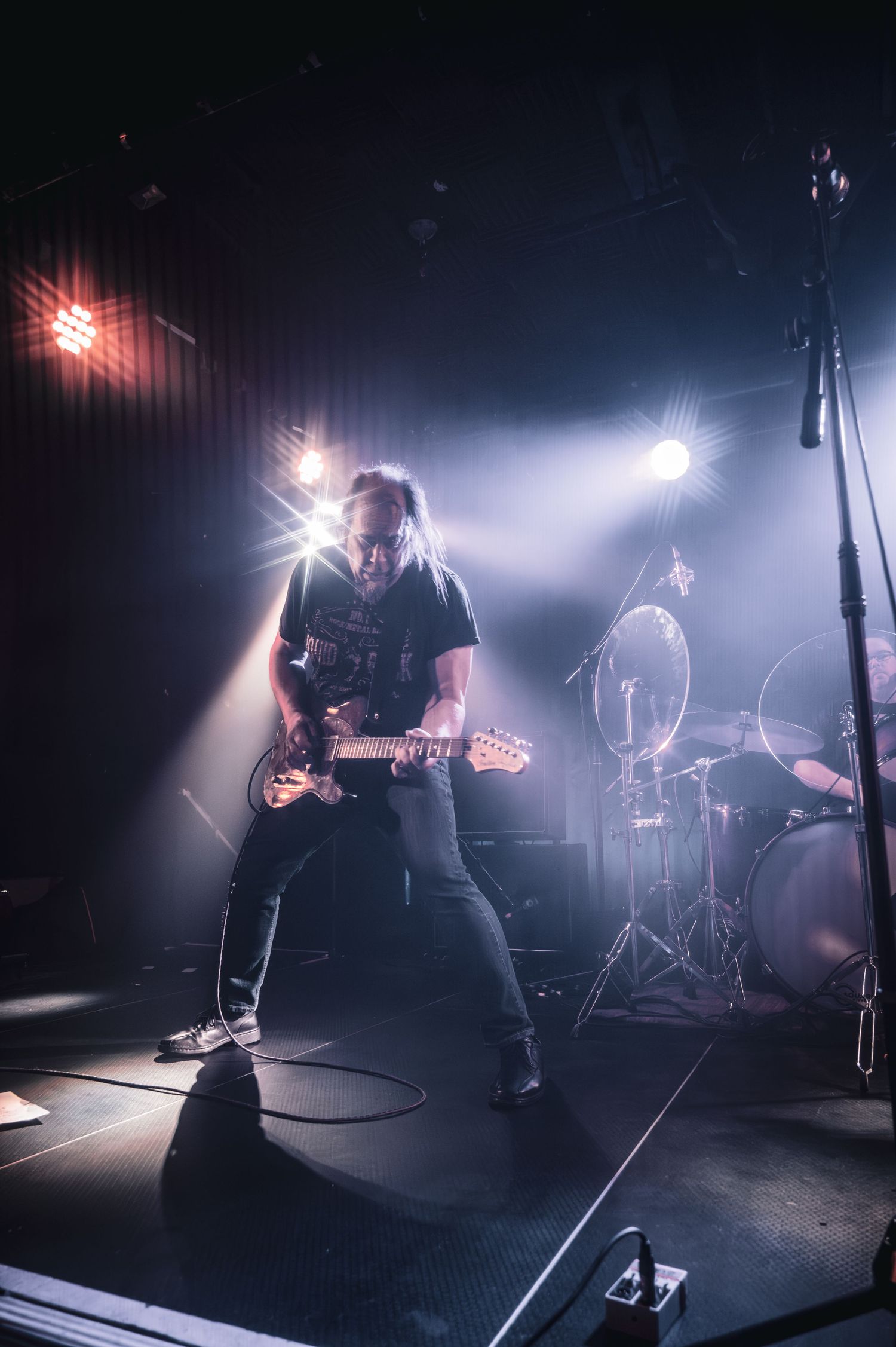Guitarist performing on stage under dramatic spotlights with drum kit visible in background at live rock concert.