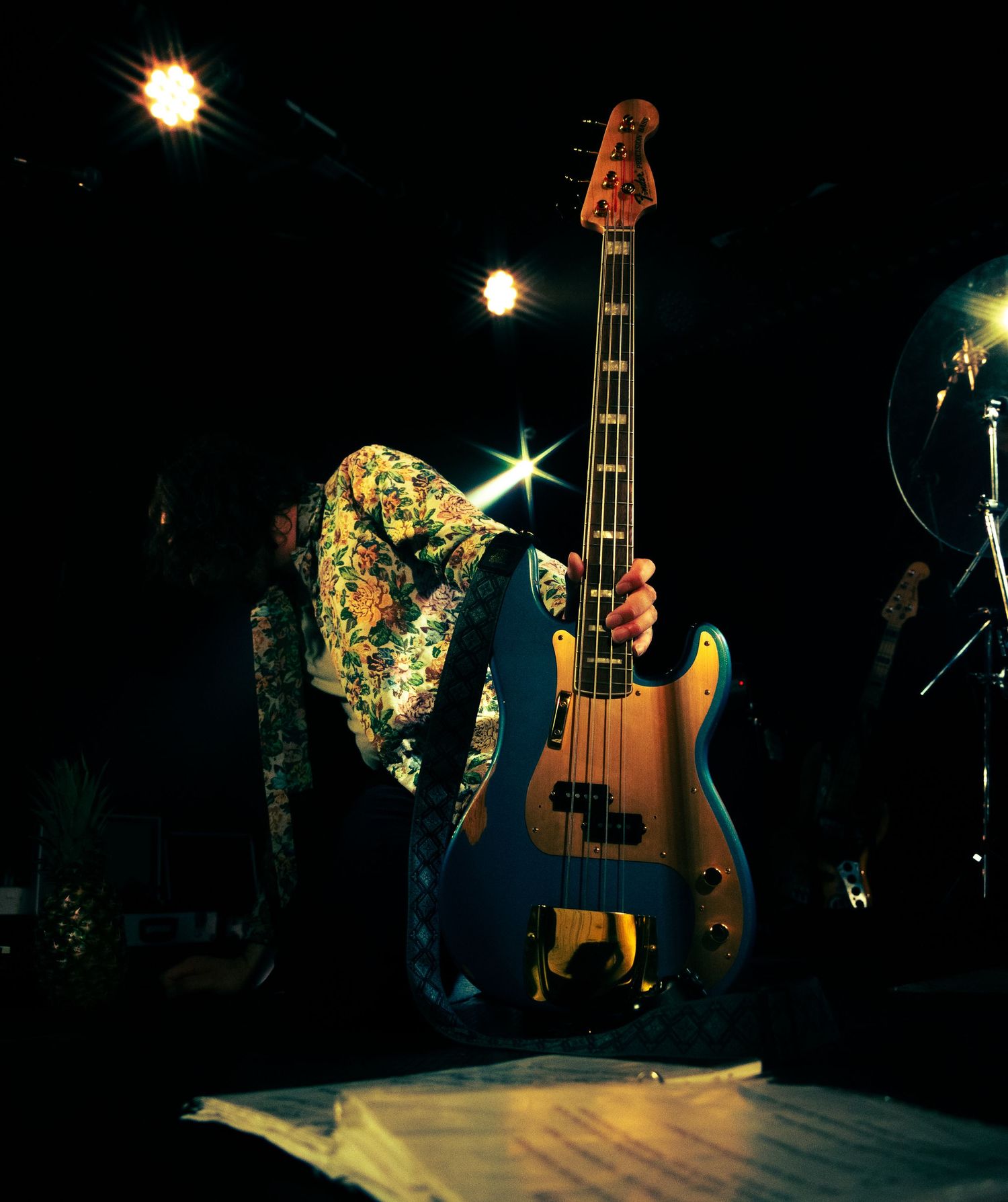 A bass guitar sits illuminated on a dark stage with dramatic lighting and sparkles creating an atmospheric concert scene.