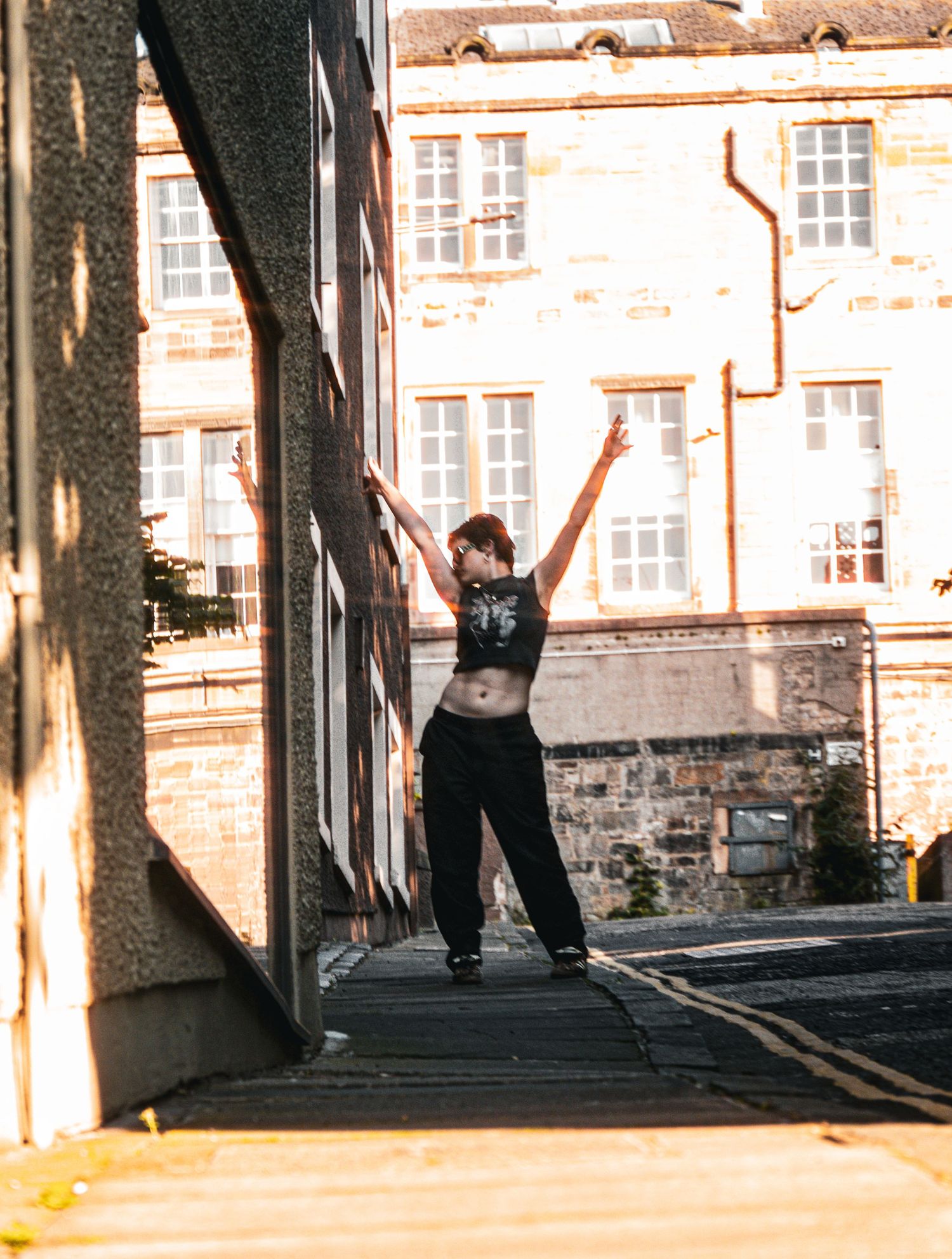 Person dancing joyfully with arms raised in a sunlit urban alleyway between historic stone buildings.