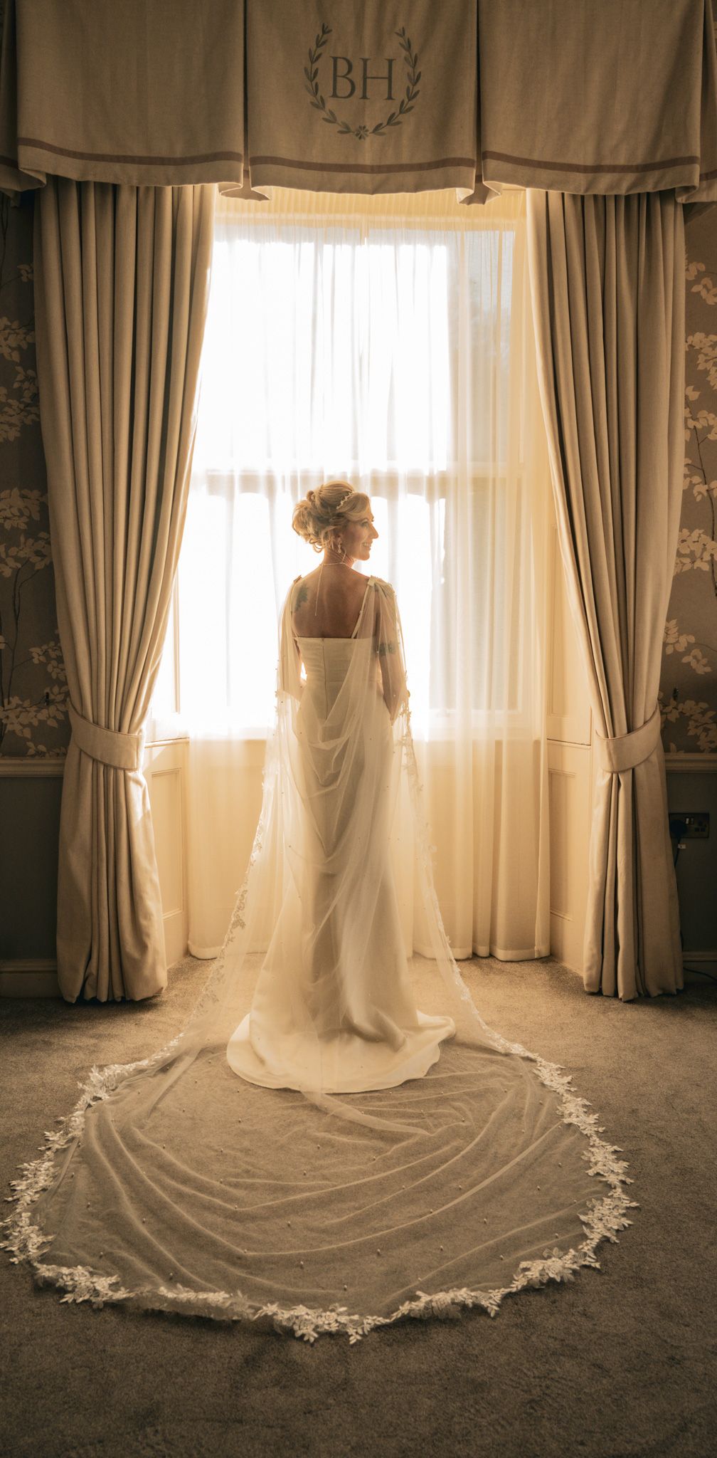 Bride in elegant white gown with lace cathedral veil stands before a sunlit window in a grand venue.