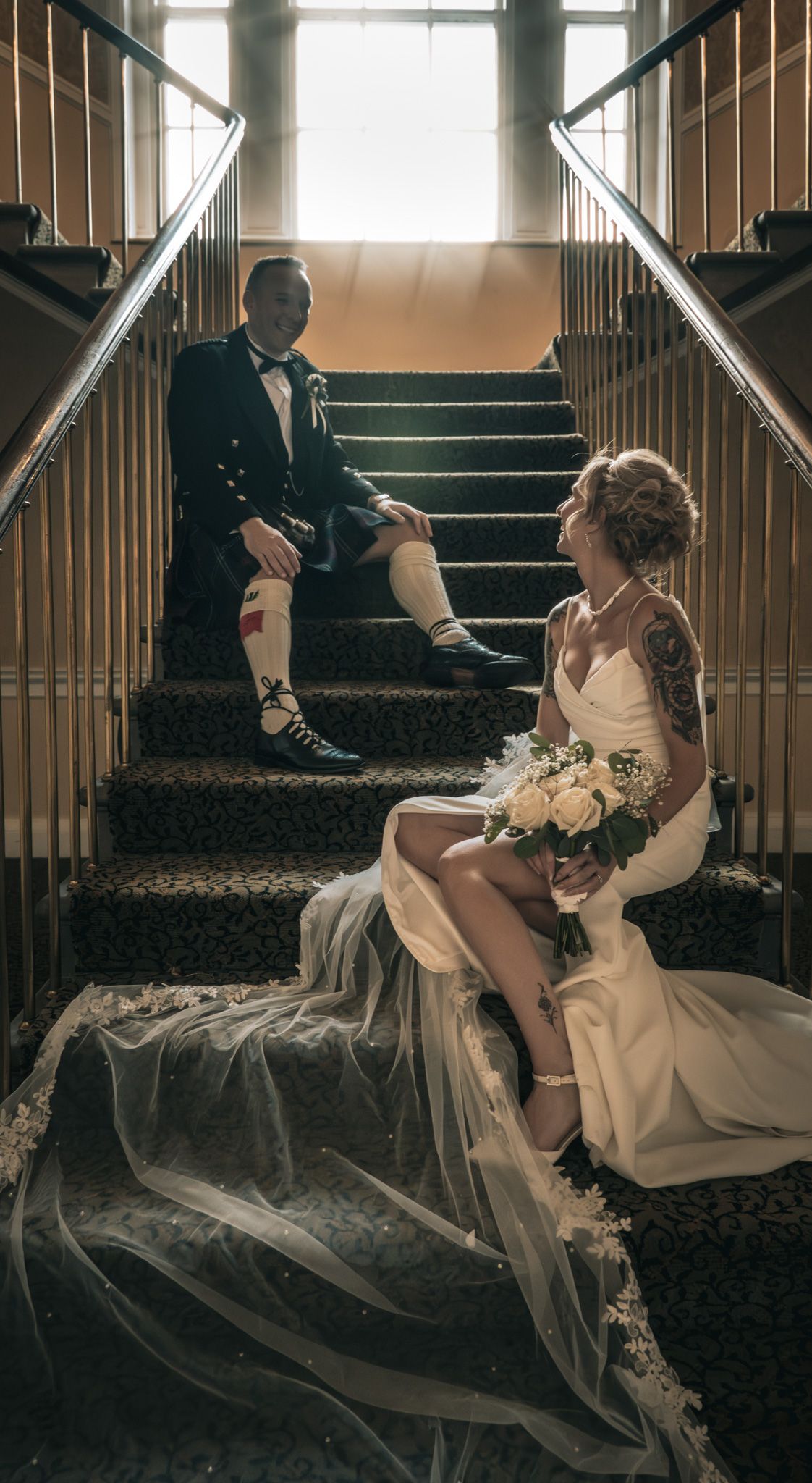 Bride and groom in Scottish kilt sit on ornate staircase, bride holding white roses bouquet with flowing veil.