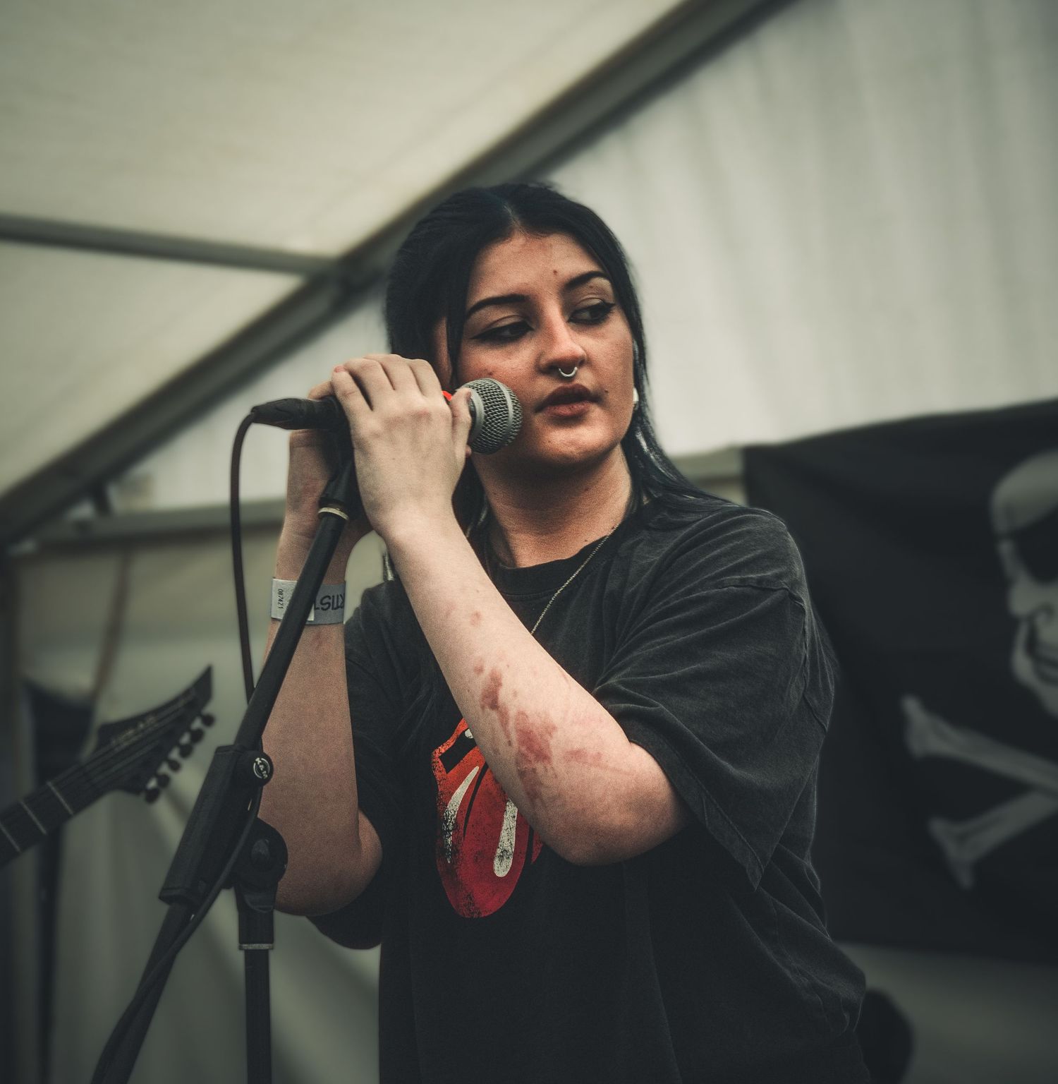Young woman with dark hair singing into a microphone on stage wearing a black t-shirt at an outdoor music event.