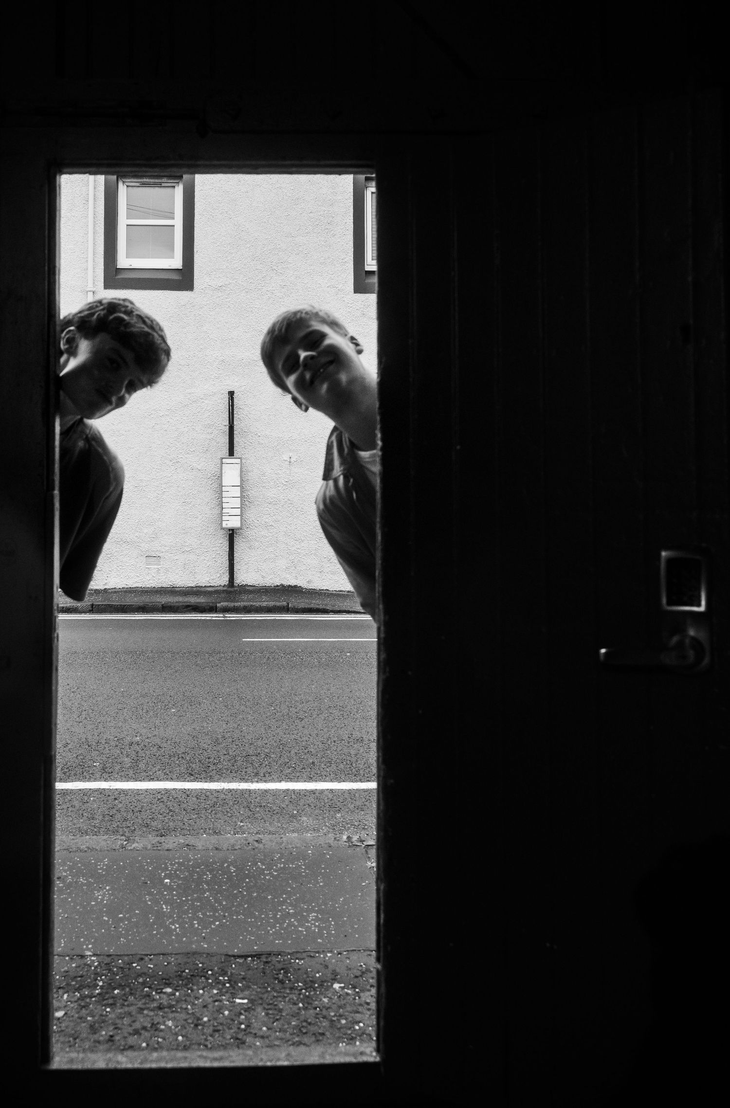 Black and white photo of two people peeking through a doorway onto a quiet street outside.