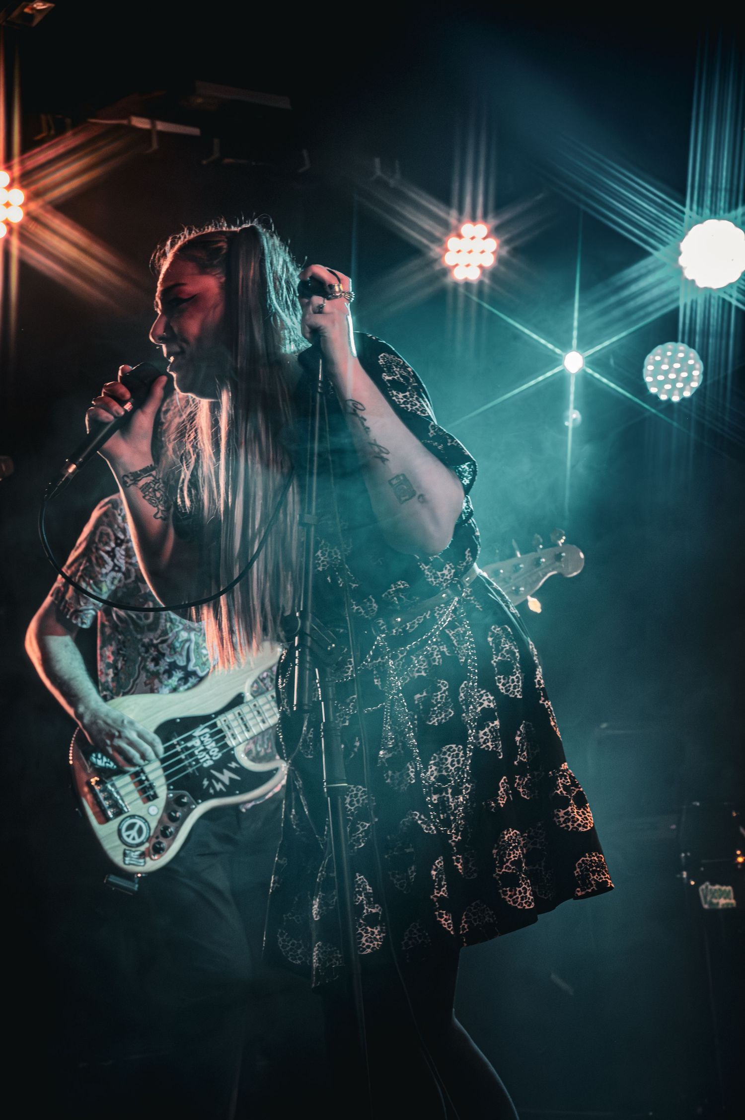 Female vocalist singing on stage with guitarist, dramatic stage lighting and smoke effects at live concert.
