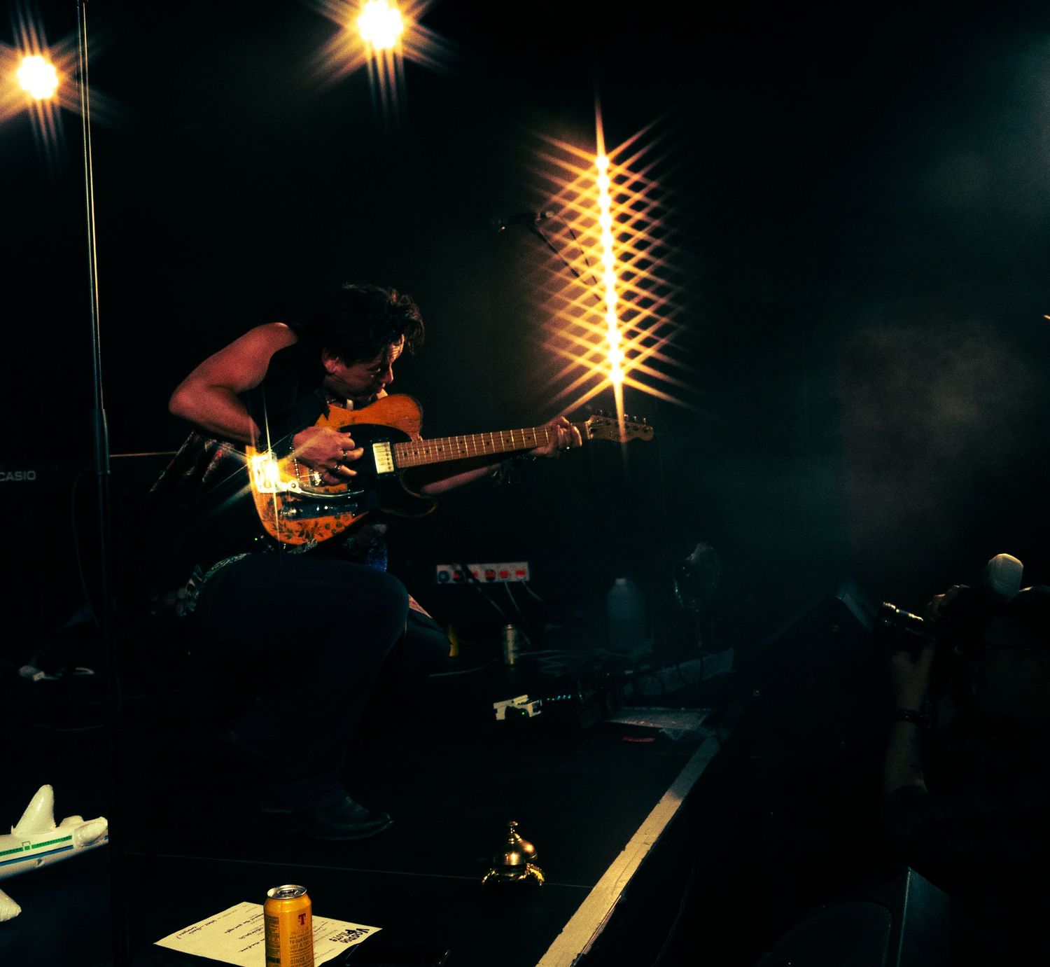 A guitarist performs energetically on stage with a bass guitar under dramatic lighting and smoke effects.