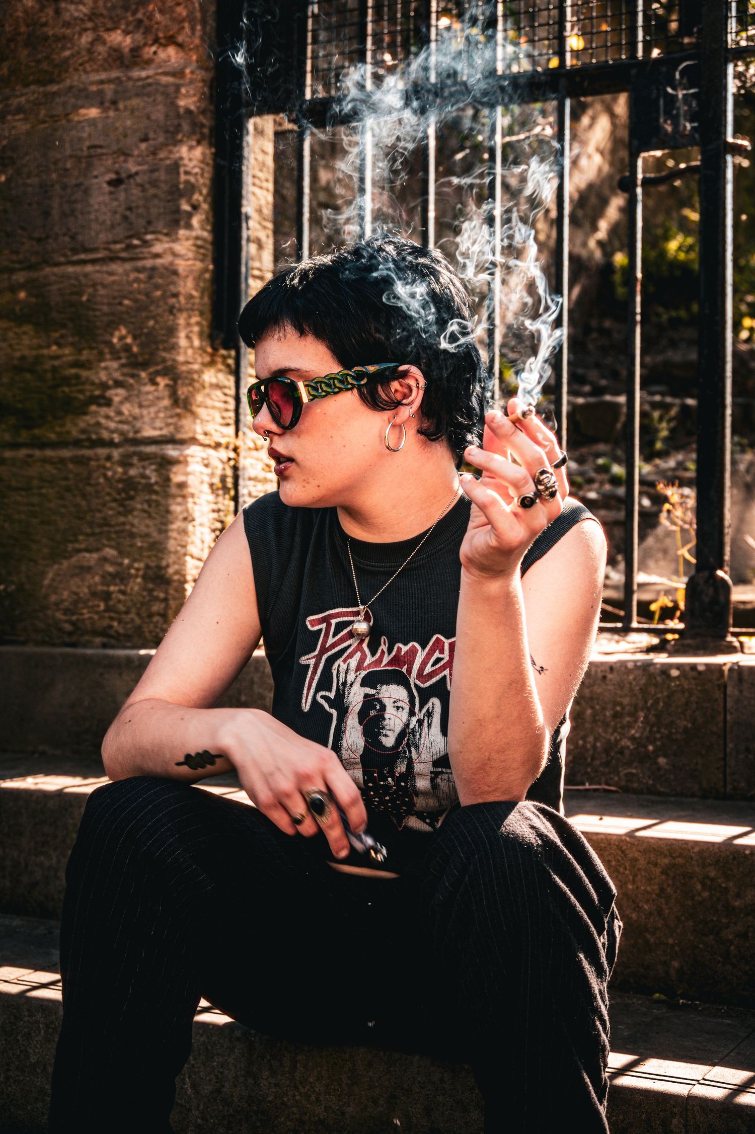 Punk-style person with short dark hair smoking, wearing sunglasses and a Prince band tee outdoors.