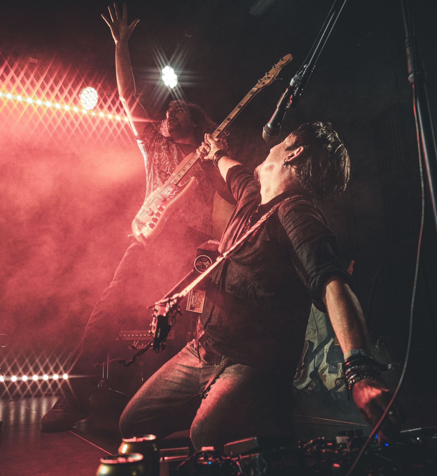 Two rock musicians perform energetically on stage under dramatic red lighting, playing electric guitars with crowd excitement.