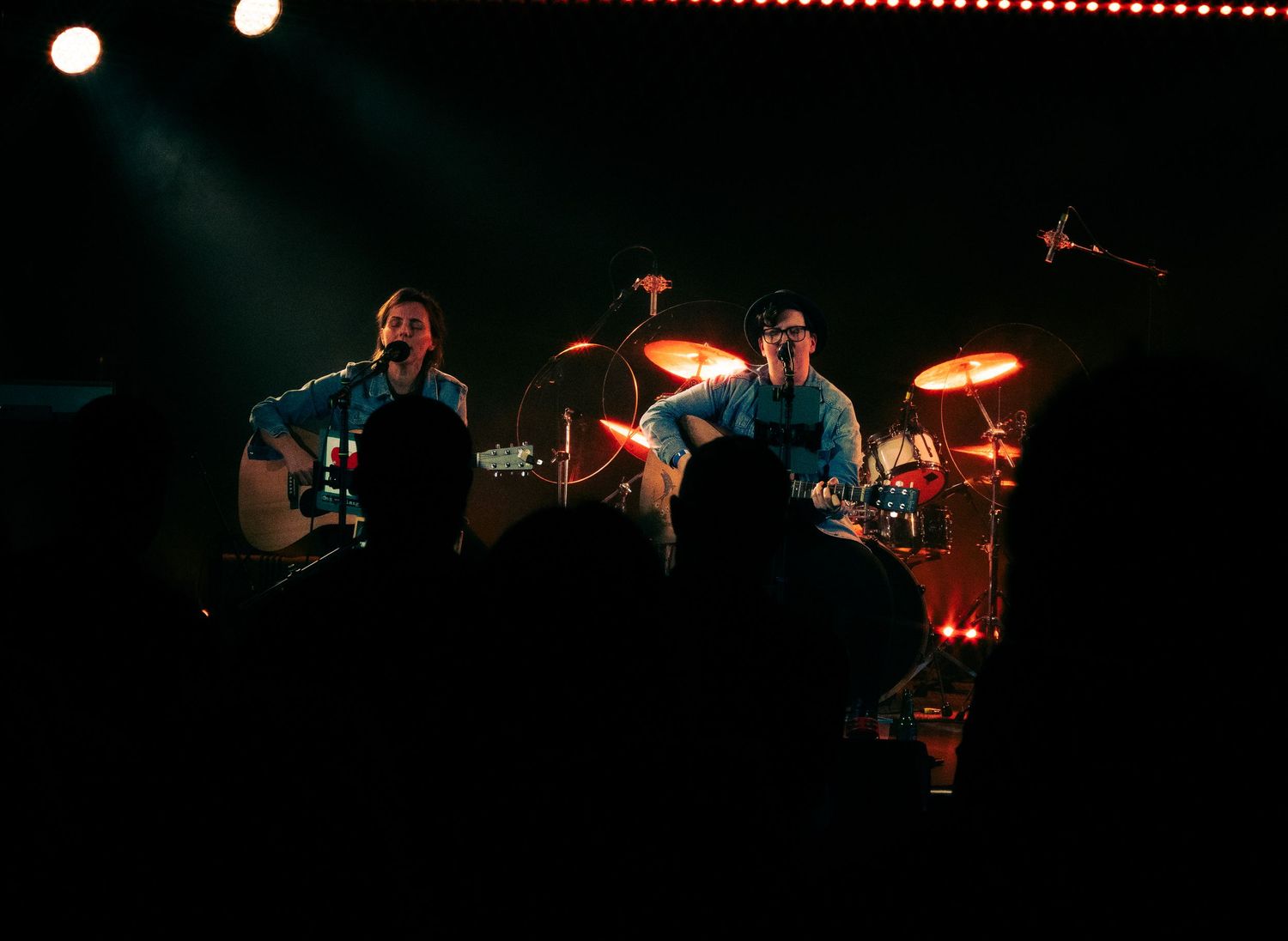 Musicians perform on stage with guitars and drums in dark lighting with red accent lights in the background.
