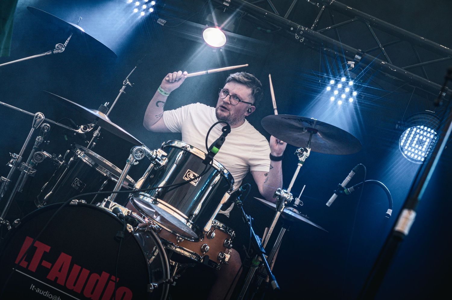 Energetic drummer performing on stage with cymbals and drum kit under blue and white stage lighting.