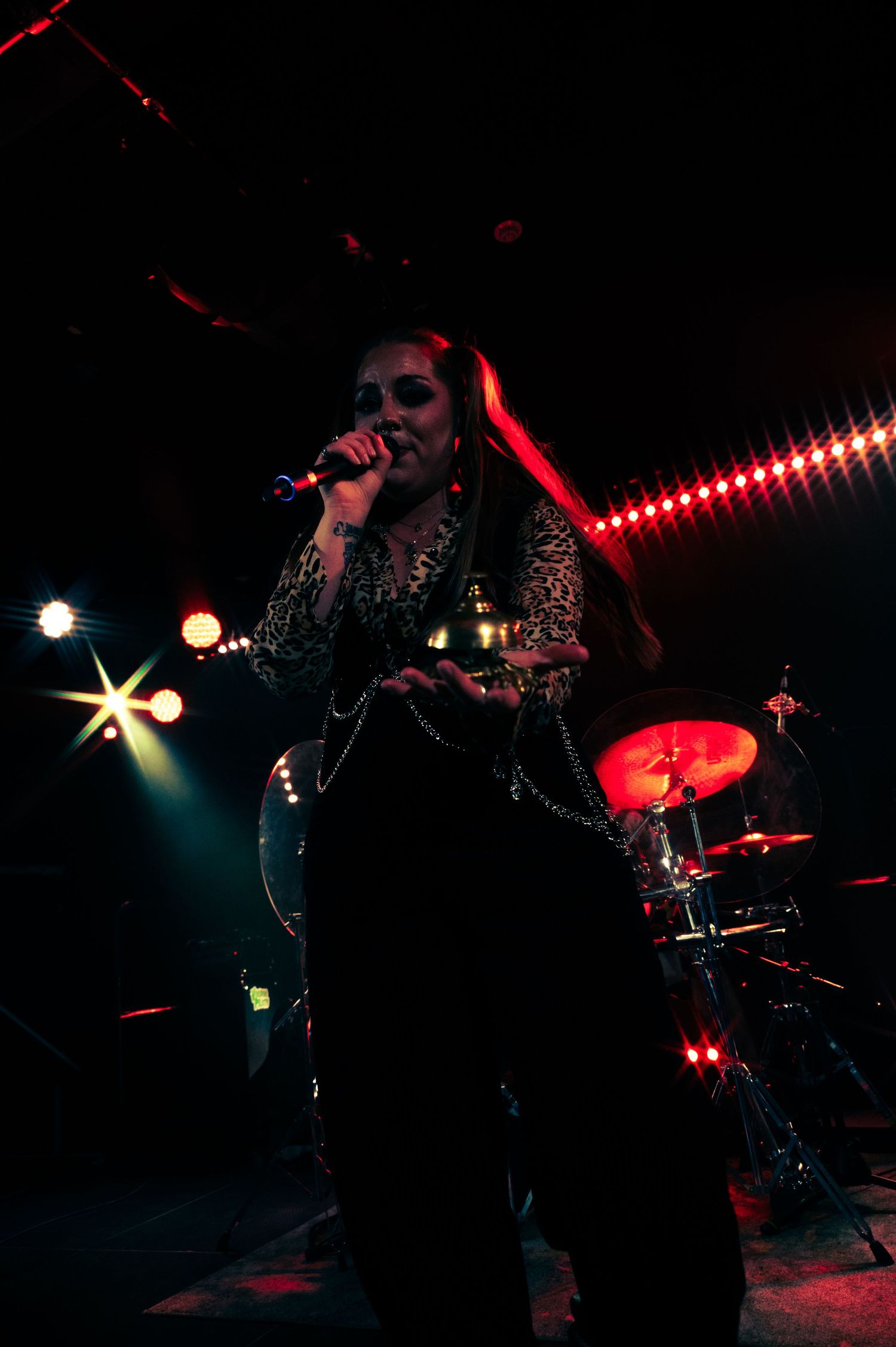 A performer singing into a microphone on a dark stage lit by dramatic red lighting and spotlights, with a drum kit visible behind.