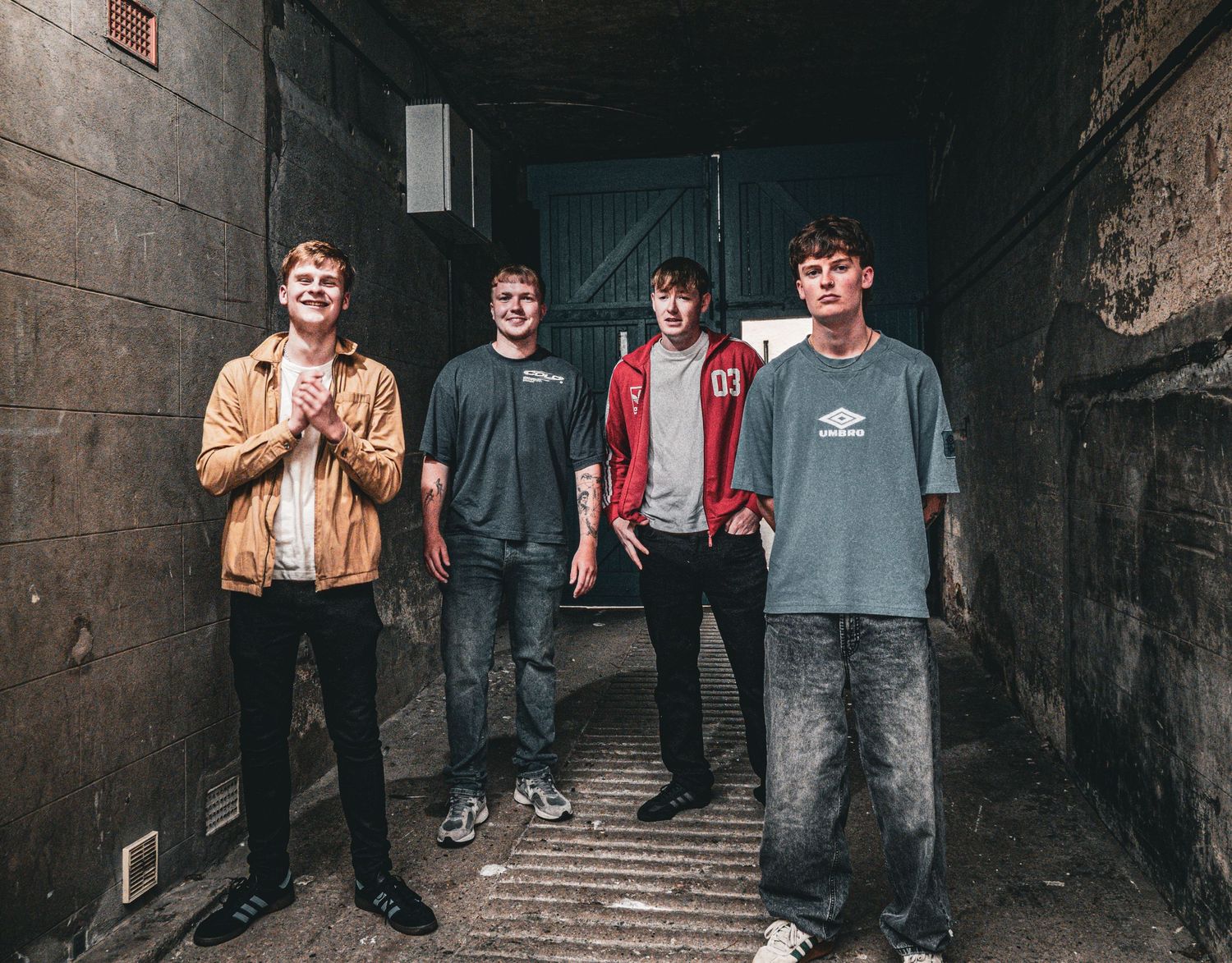 Four young men stand in an urban alleyway wearing casual streetwear including a yellow jacket and red hoodie.