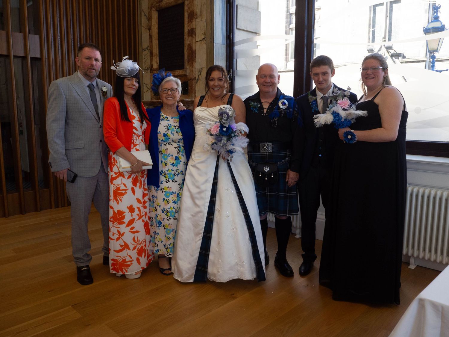 Wedding party of seven people posing together, bride in white and blue gown, groom in Scottish kilt, formal attire.