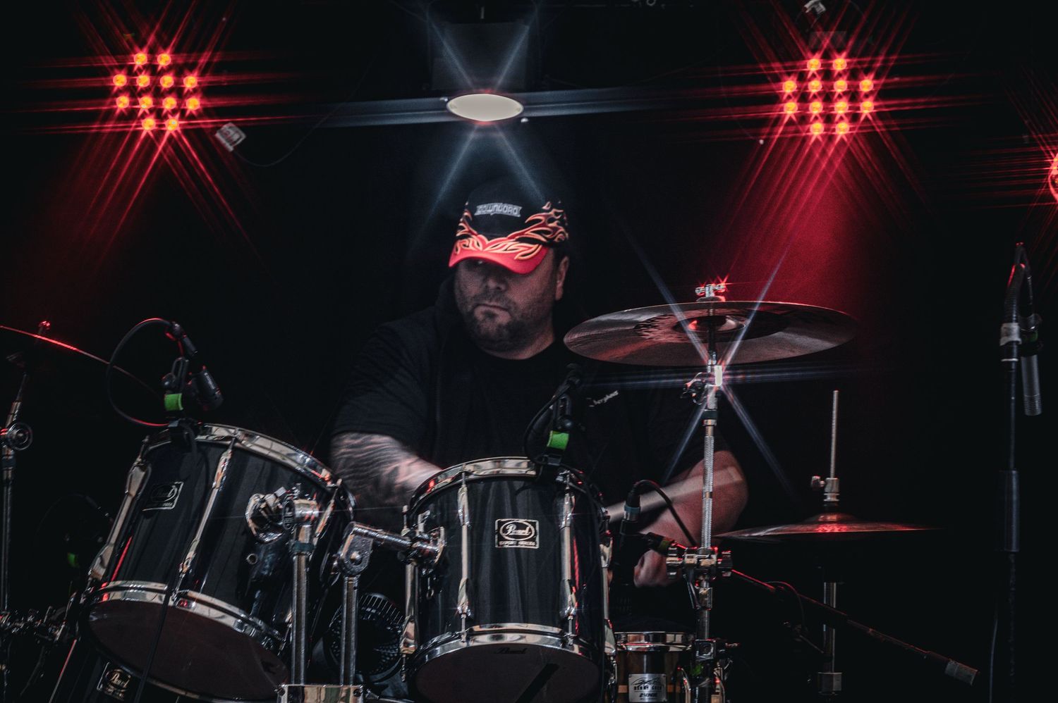 Male drummer playing drum kit on stage with red stage lighting behind him.