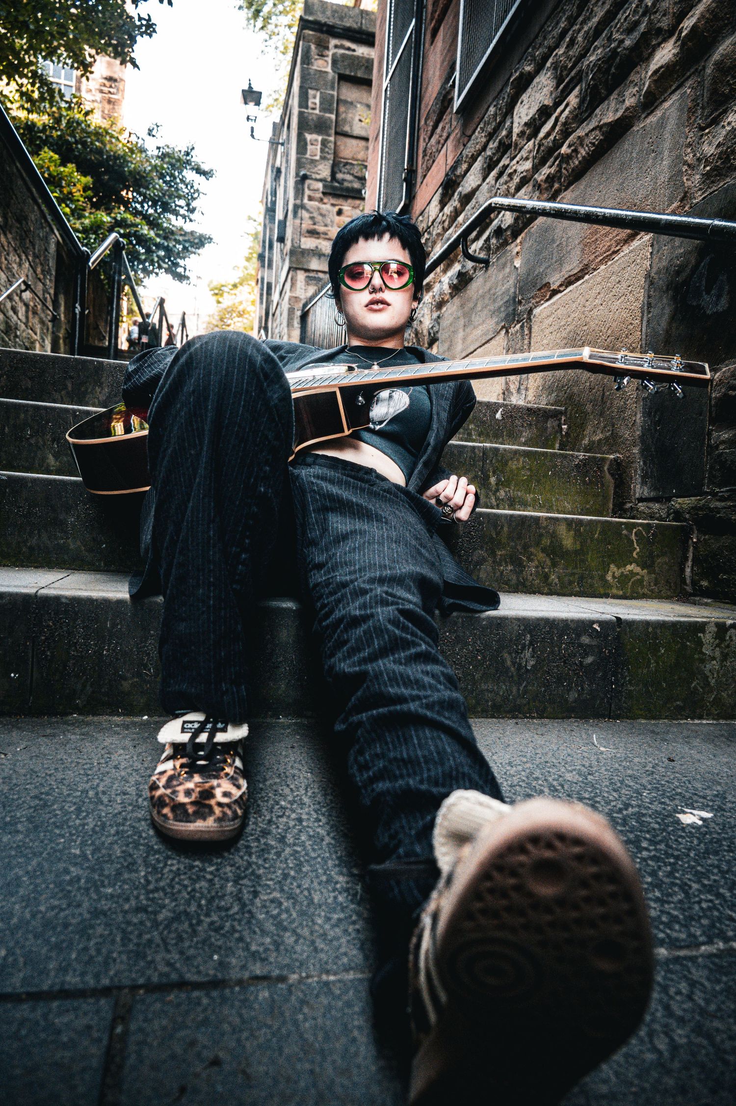 Musician in pinstripe suit and red sunglasses lounging on stone steps with acoustic guitar.