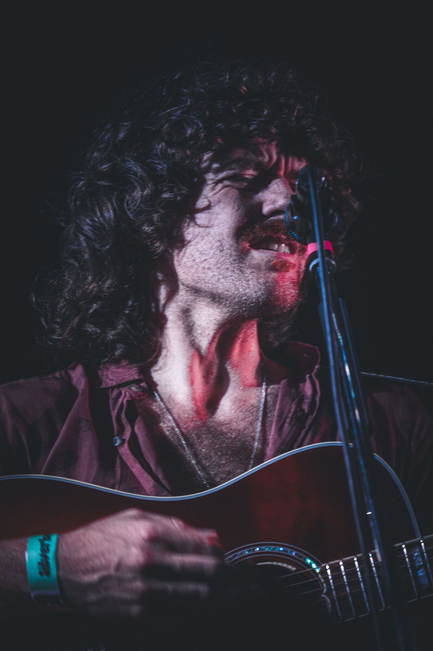 Curly-haired male guitarist singing passionately into microphone under red stage lighting.