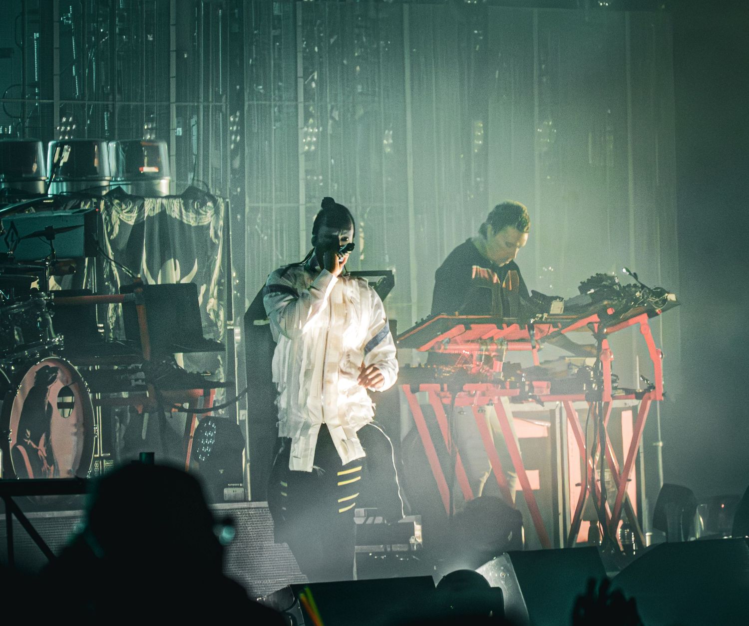 Two performers on a dramatic fog-filled stage, one in white outfit singing, another DJing at a red illuminated console.