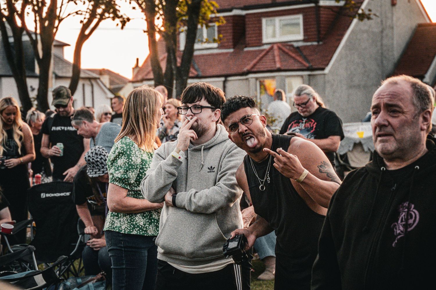 Group of people gathered outdoors at a backyard event, with two men playfully posing in the foreground at dusk.