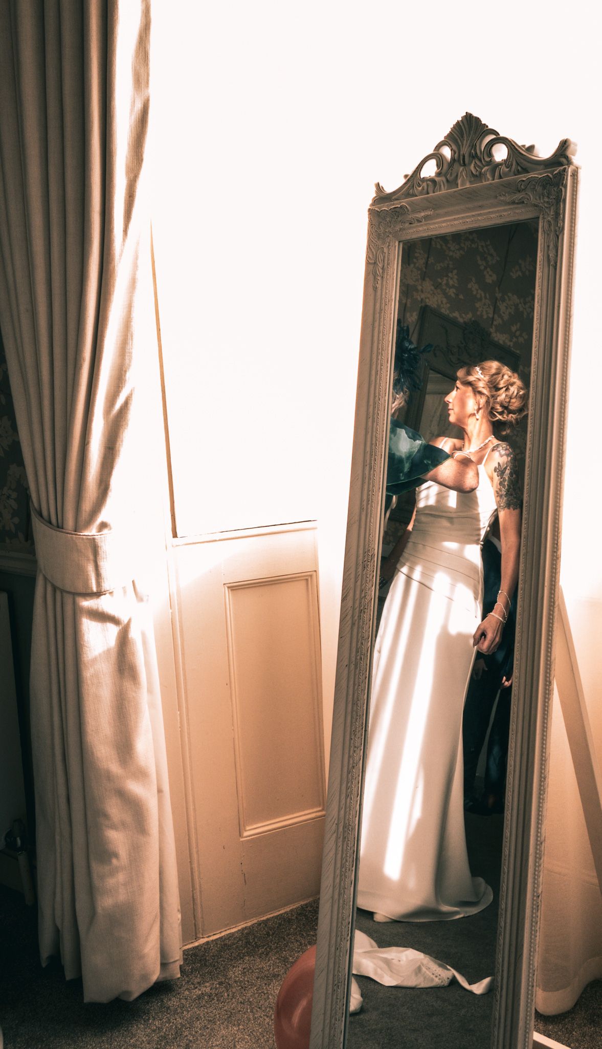 Bride in white wedding gown admiring her reflection in an ornate full-length mirror beside draped curtains.